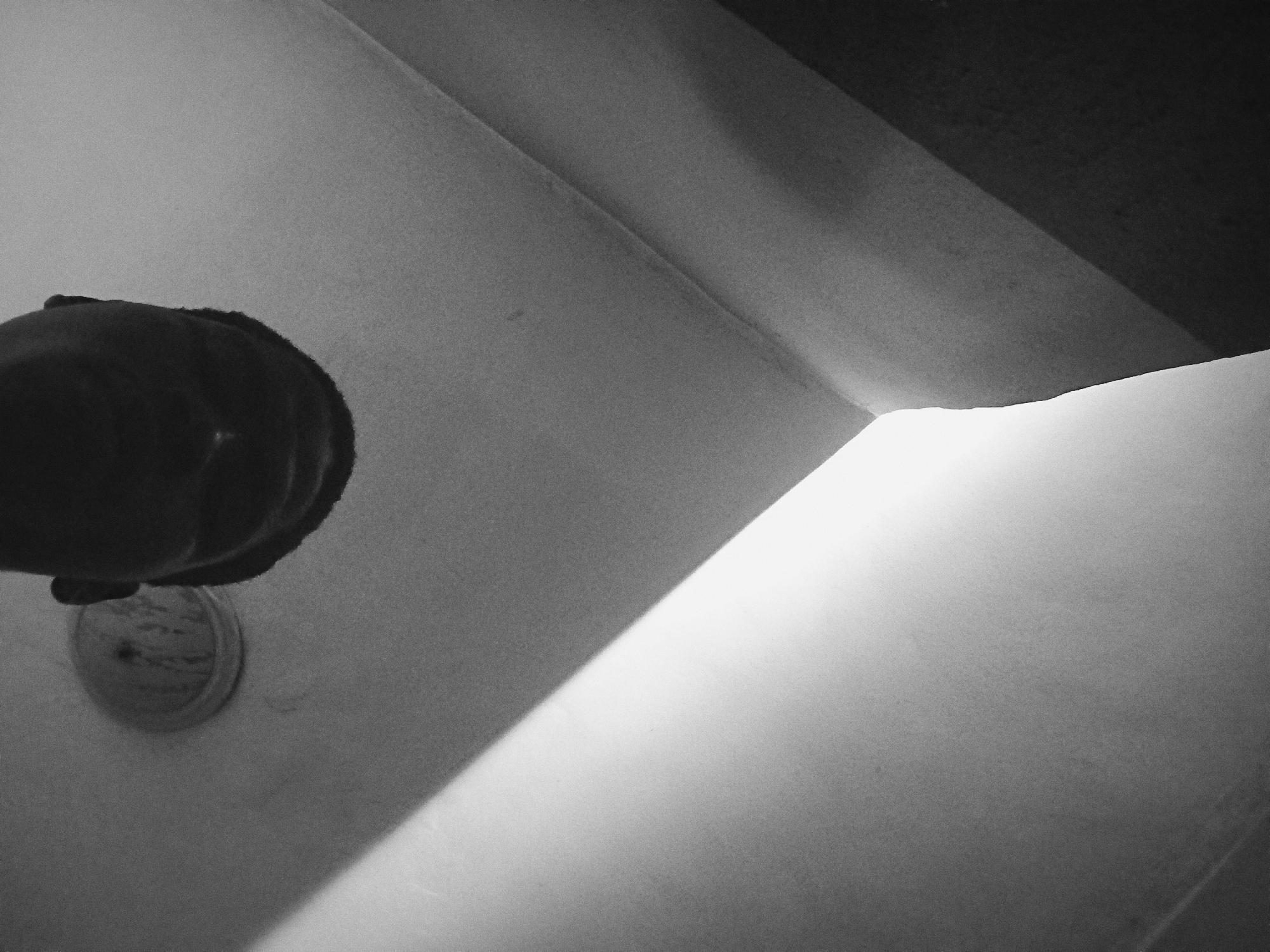 A low-angle, black and white photograph shows a toilet. To the left is a dark, round toilet seat and a toilet bowl fixture, both partially visible. A bright light reflects off a white surface in the lower-right corner, obscuring details. The upper-right corner is dark. The location, Nkpor, Nigeria, is not discernable from the image.