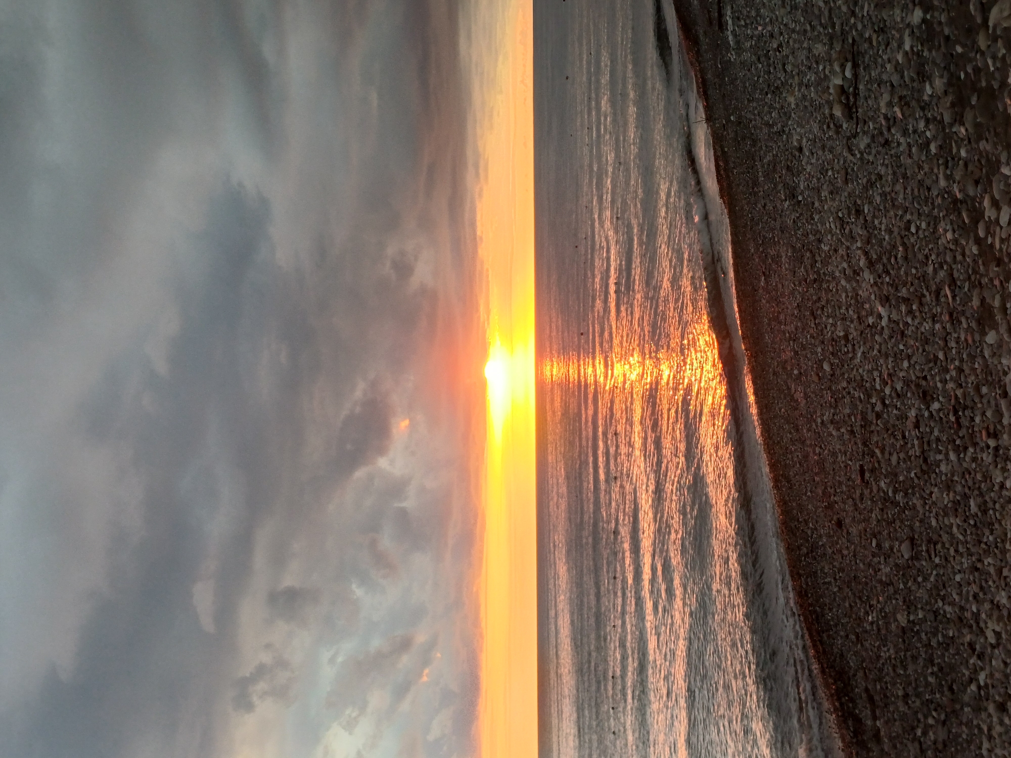 The image depicts a sunset over the Black Sea in Sochi, Russia. The sky is filled with a mix of dark gray and orange clouds, with the setting sun casting a bright orange glow on the horizon. The water is calm, with gentle waves lapping at the pebbled shore. There are no people or boats visible in the image. The scene is serene and tranquil, with the natural beauty of the sunset dominating the frame. The foreground is composed of a dark, rocky beach, leading into the calm sea. The overall impression is one of peacefulness and the end of a day.