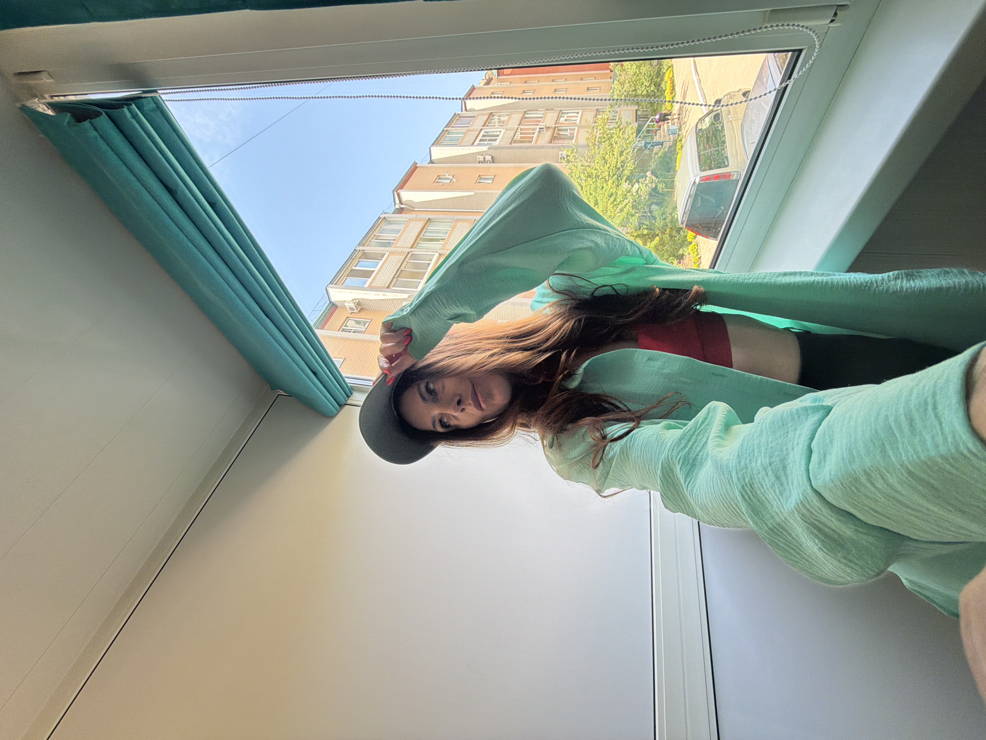 A woman with long brown hair and a grey fedora is positioned near a window. She wears a red top and a light green, textured garment draped around her. She is looking at the camera.
Outside the window, buildings and greenery are visible under a bright sky. A light-colored vehicle is parked on the street. The location is Birobidzhan, Russia based on the architectural style of the buildings and the local environment.
The interior appears to be a room with a white wall and a blue-green curtain or blind next to the window. The photo is taken from a low angle, making the woman and the window the primary subjects.