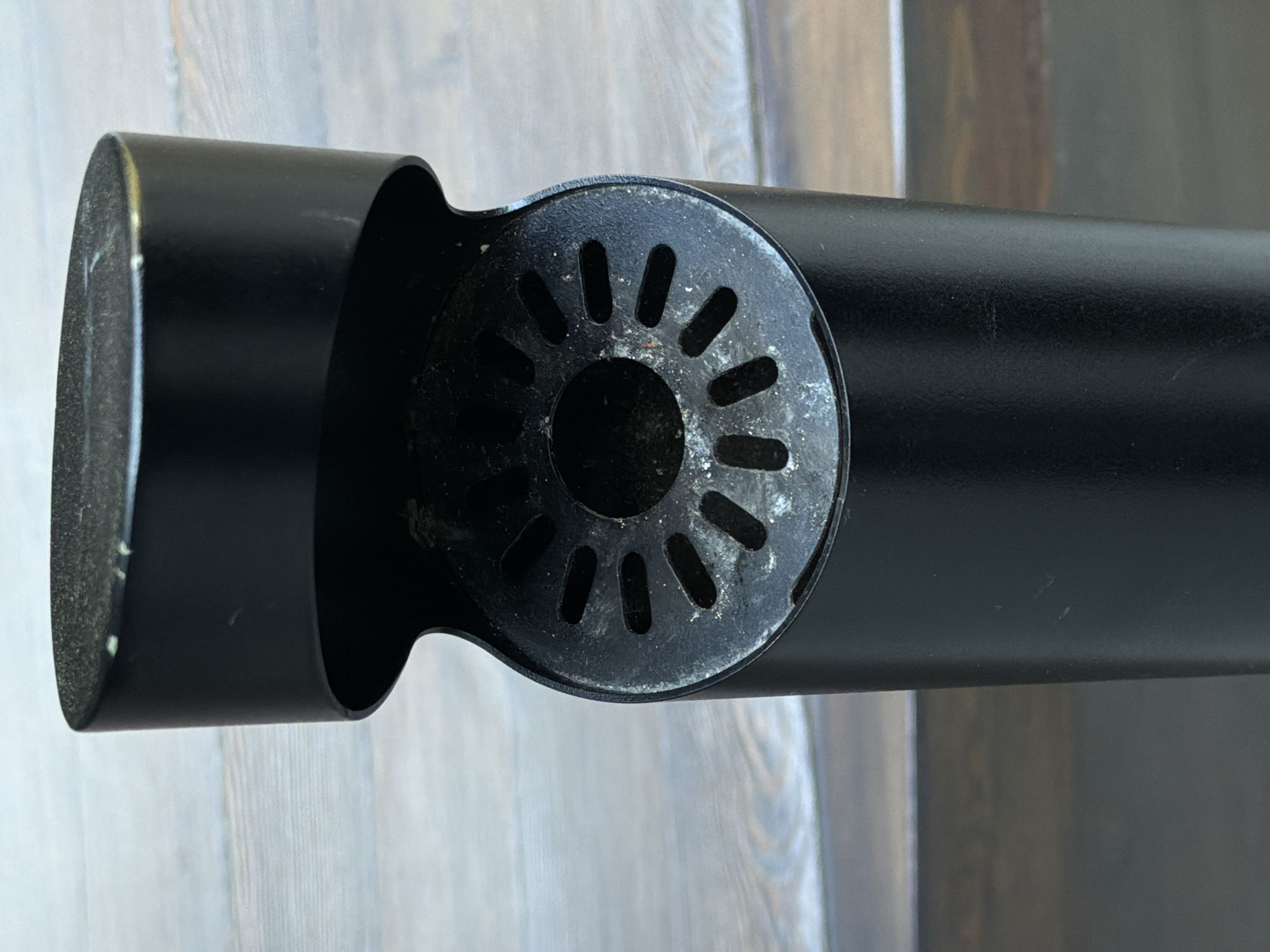 The image presents a close-up, horizontal view of a black, cylindrical object, likely a metal tube or pipe, positioned against a blurred light-colored wooden background. The object has a matte, slightly textured surface.
A prominent feature is a circular, perforated disc or grate embedded within a concave cut-out on one side of the cylinder. This disc, also black, displays a pattern of radiating, elongated slots surrounding a central circular hole. The surface of this perforated component, along with parts of the cylindrical object, exhibits visible specks of white-ish residue or dust, suggesting use or exposure. The far end of the cylinder also shows similar light-colored markings along its edge.
The background is out of focus, revealing the natural grain and light tones of a wooden surface, possibly a table or floor. The lighting appears soft and even, characteristic of an indoor setting. No people are visible, nor is any specific activity or event depicted; the image is a static shot focusing on the object's details. There is no visible text. The provided crops offer closer views of the object's texture, the intricate design of the perforated disc, and the subtle details of the background wood grain.