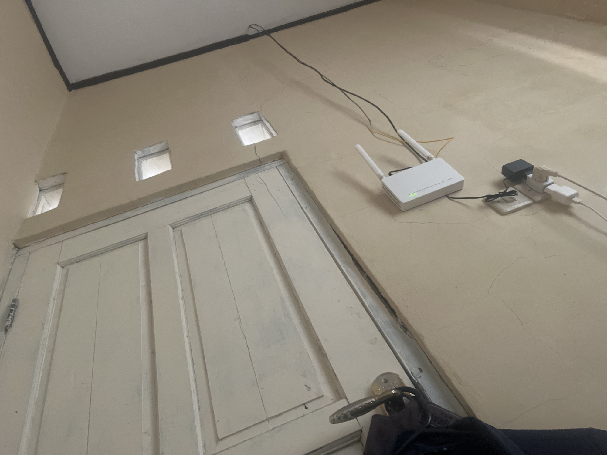 This image shows a close-up of a wall in Cirahayu, Indonesia, with a white door, a WiFi router, and electrical outlets. The wall is painted a light beige color and has visible cracks. To the left, a white door with horizontal panels is slightly ajar, revealing a dark object, possibly a piece of clothing. A metallic door handle is visible on the door.
Above the door, there are three small square openings in the wall. To the right of the door, a white WiFi router with two antennas is plugged into a power strip. The router has a green indicator light. Connected to the power strip are three electrical plugs, one of which is a black adapter. Black and yellow wires are connected to the router and extend upwards. The overall lighting suggests an indoor setting, possibly a room or hallway.