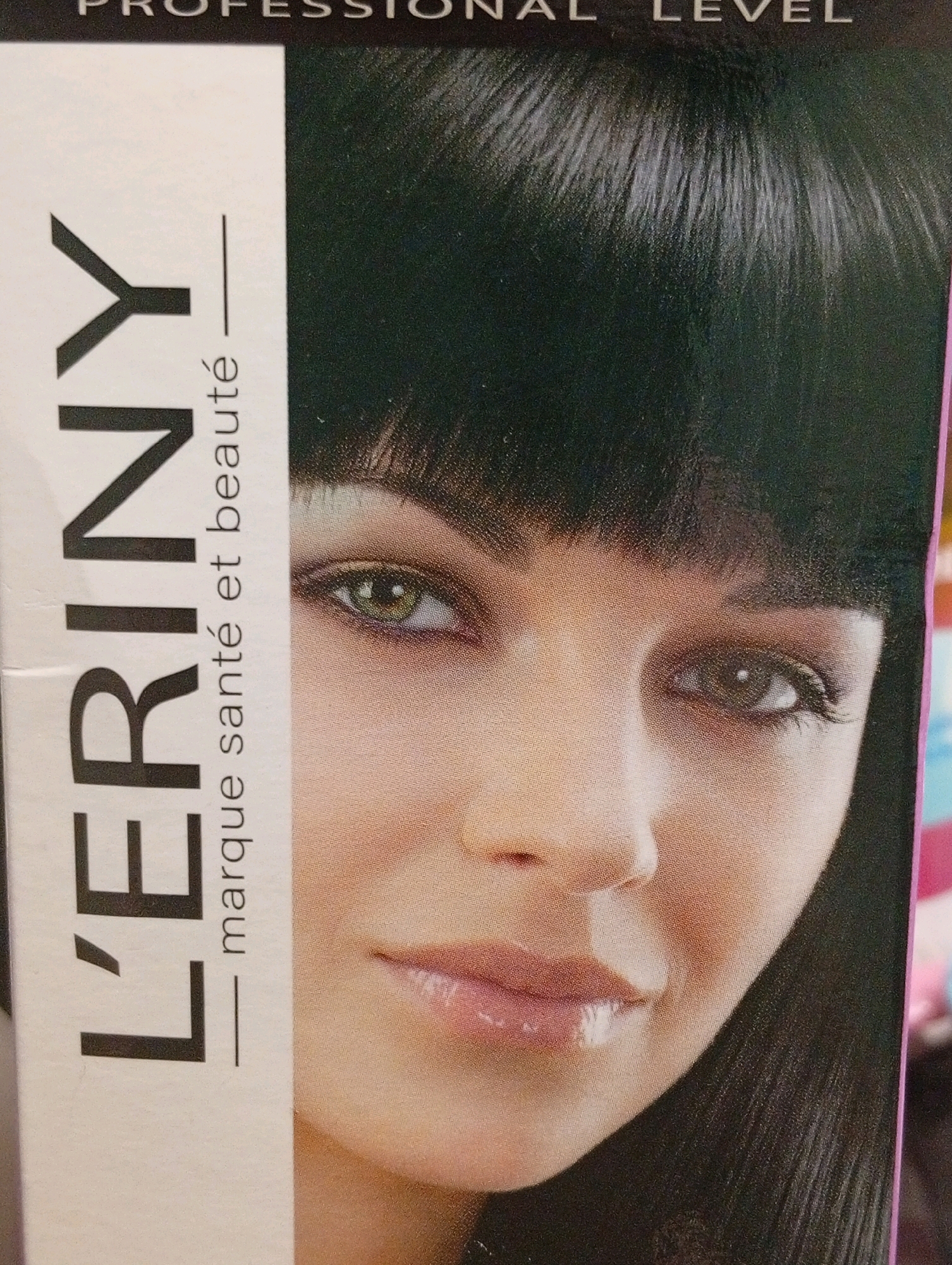The image is a close-up shot of a box of hair dye featuring a woman's face. The woman has black hair and green eyes. Text on the box reads "PROFESSIONAL LEVEL" at the top and "L'ERINY" vertically along the left side, with "marque santé et beauté" below it. The woman's makeup includes eyeshadow and lip gloss. The background is dark, suggesting the focus is on the product packaging and the woman's face. There is no indication of a specific city or location.