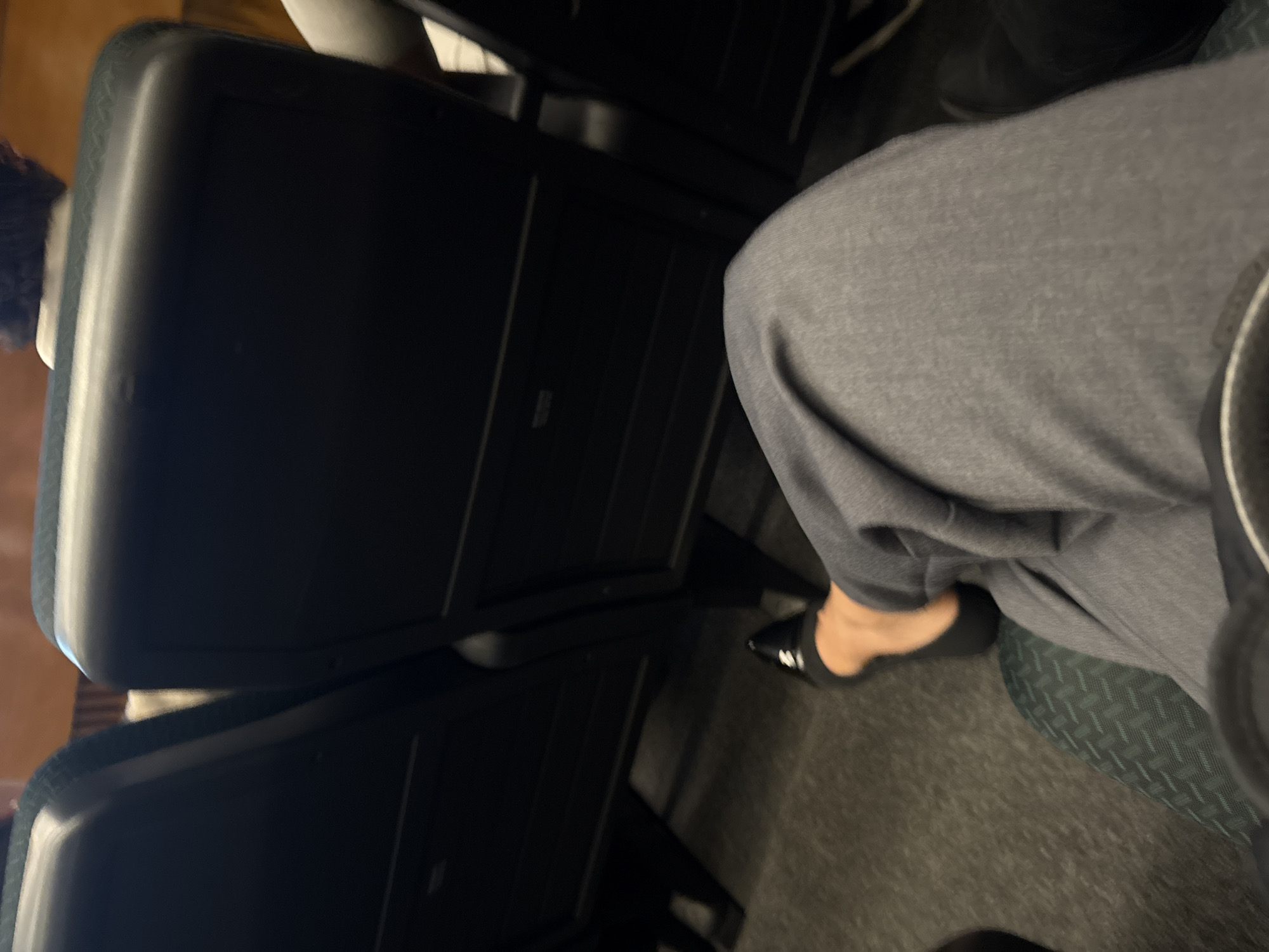 The image shows an airplane seat. A person wearing gray pants and black shoes is seated. The plane appears to be on the ground, with the floor visible. The location in the city of San Andrés Cholula, Mexico is not discernible from this image.