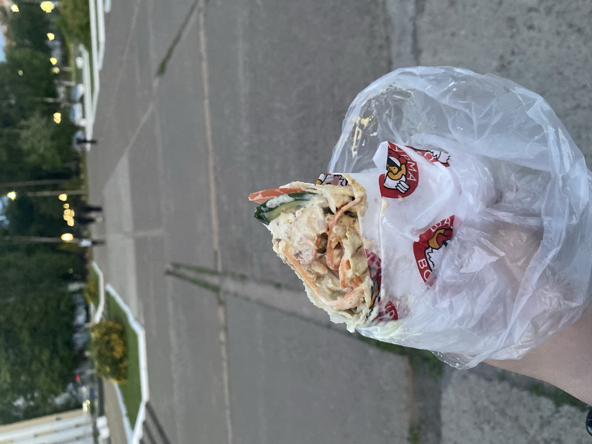 A person's hand holds a shawarma wrapped in paper and a plastic bag. The shawarma is partially unwrapped, revealing layers of filling including shredded carrots, cucumber slices, and a creamy sauce. The paper wrap has red and white branding with cartoon-like images. The background is a blurred outdoor urban setting, possibly a park or plaza, with trees, streetlights, and a few people walking in the distance. The lighting suggests it is late afternoon or early evening.
