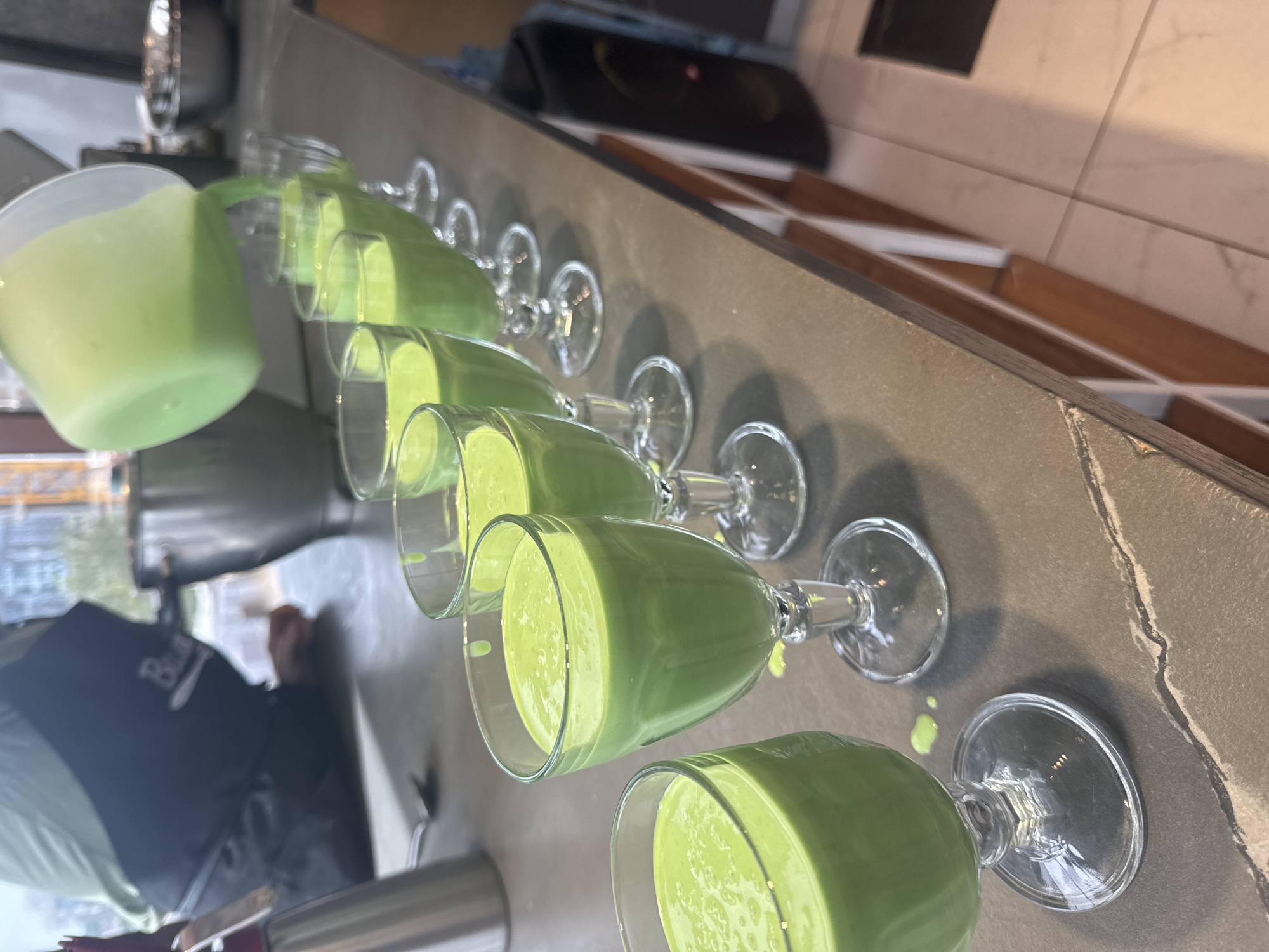A person is pouring a green liquid into a row of stemmed glasses, presumably at a counter. The liquid is opaque and light green, suggesting a smoothie or similar drink. The glasses are lined up on a grey countertop, with a few droplets of the liquid spilled onto the surface. The background shows a window looking out onto a city scene, but the location is difficult to pinpoint with certainty. However, the query specifically states that the city is Izhevsk, Russia. The interior appears to be a kitchen or bar setting. A person is visible, wearing what looks like a dark jacket or apron. The image focuses on the process of pouring the drinks and the arrangement of glasses.
