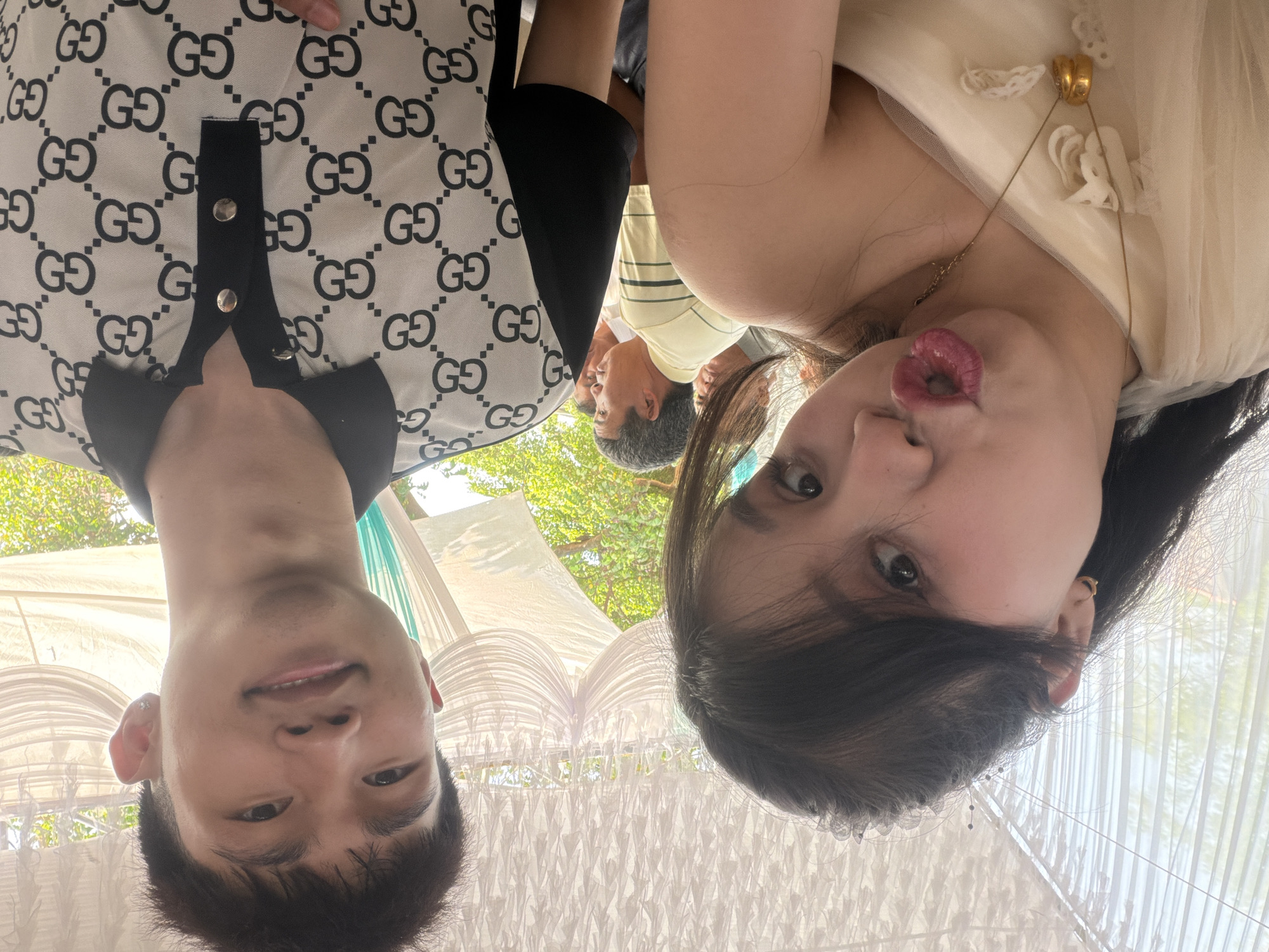 Two young adults pose cheek to cheek under a white tent-like structure, presumably at a wedding venue. The male, on the left, wears a black and white patterned shirt and smiles at the camera. The female, on the right, puckers her lips and wears a cream-colored dress with a gold necklace. An older man in a pale striped shirt stands in the background. The location is likely the city of Bình An, Vietnam.