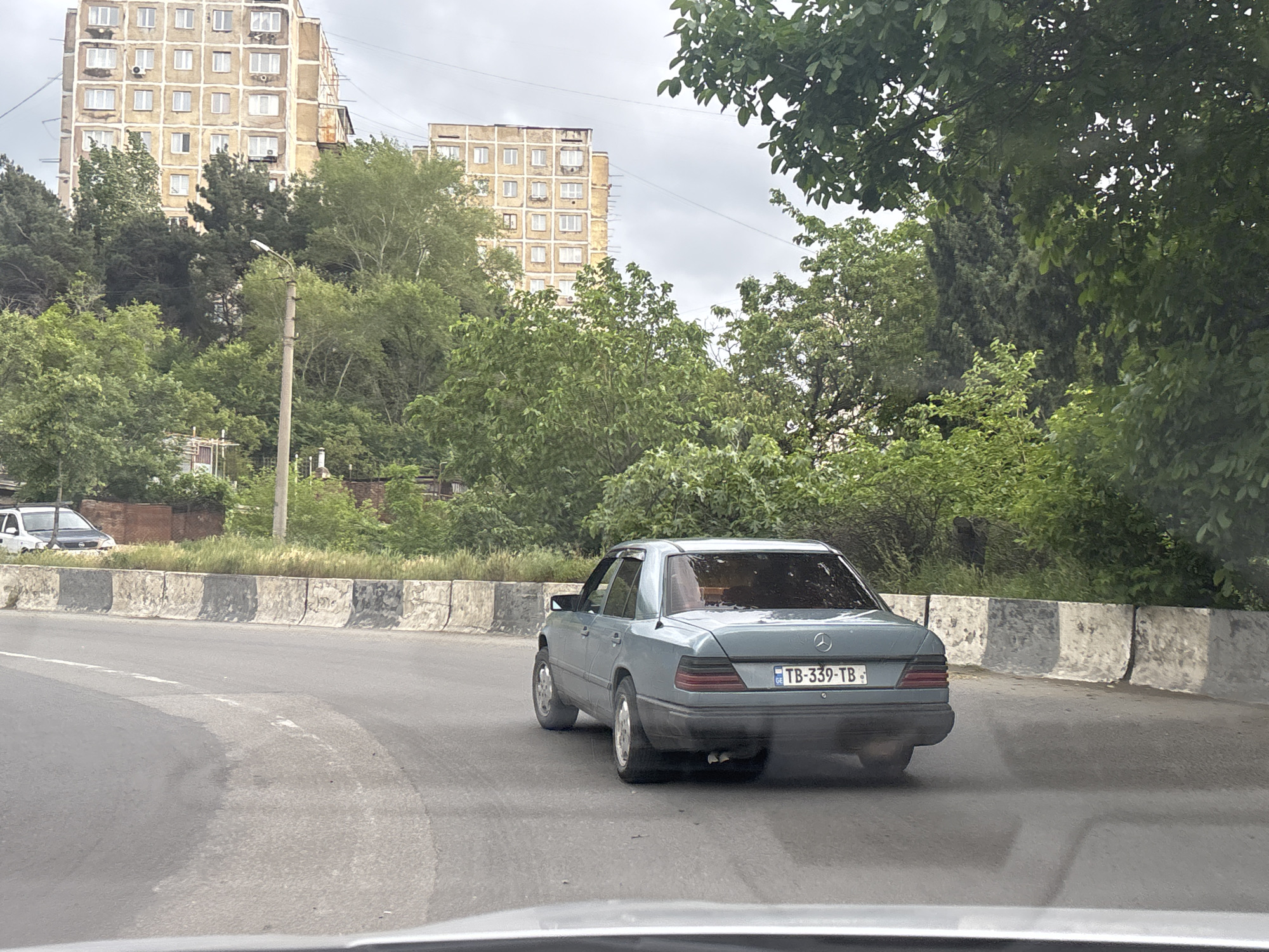 A photograph taken from inside a vehicle depicts a grey Mercedes-Benz sedan driving on a paved road in Tbilisi, Georgia. The car's license plate reads "TB-339-TB." A concrete barrier lines the edge of the road, separating it from green vegetation and trees. In the background, there are multiple-story residential buildings. The weather appears overcast. There are no visible people.