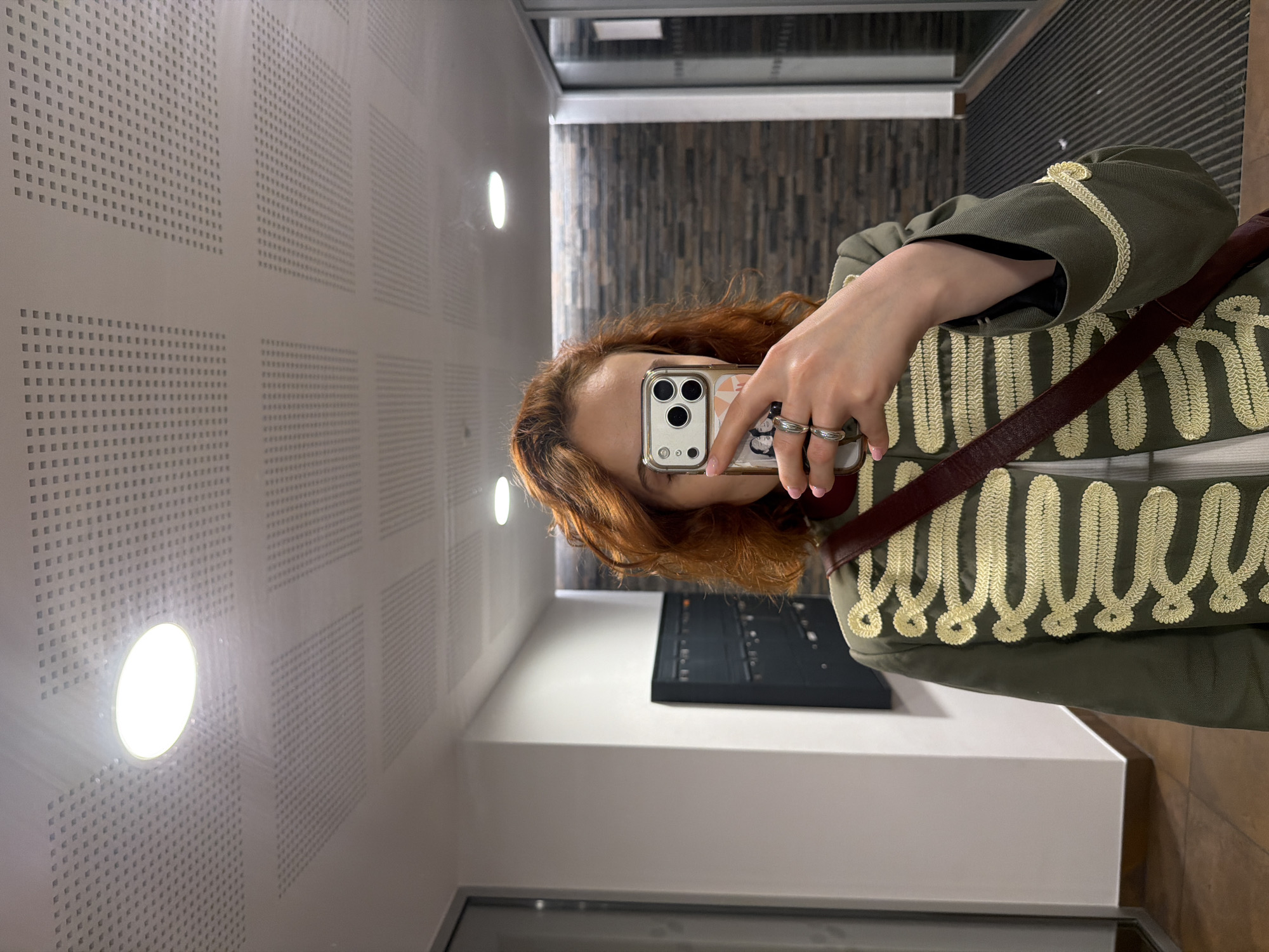 In a dimly lit indoor corridor, a woman with auburn hair takes a selfie using her phone. She is wearing an olive green jacket adorned with ornate gold trim and a maroon strap across her chest. Her left hand, adorned with two silver rings, is visible holding the phone. Behind her, a wall with a textured dark brick pattern runs horizontally, and a modern ceiling with recessed lighting panels is visible above. To her left, a white wall and a dark set of mailboxes are present. The floor is tiled with a pattern of brown squares. The scene appears to be an interior hallway, possibly in a residential building or a public space in Strasbourg, France.