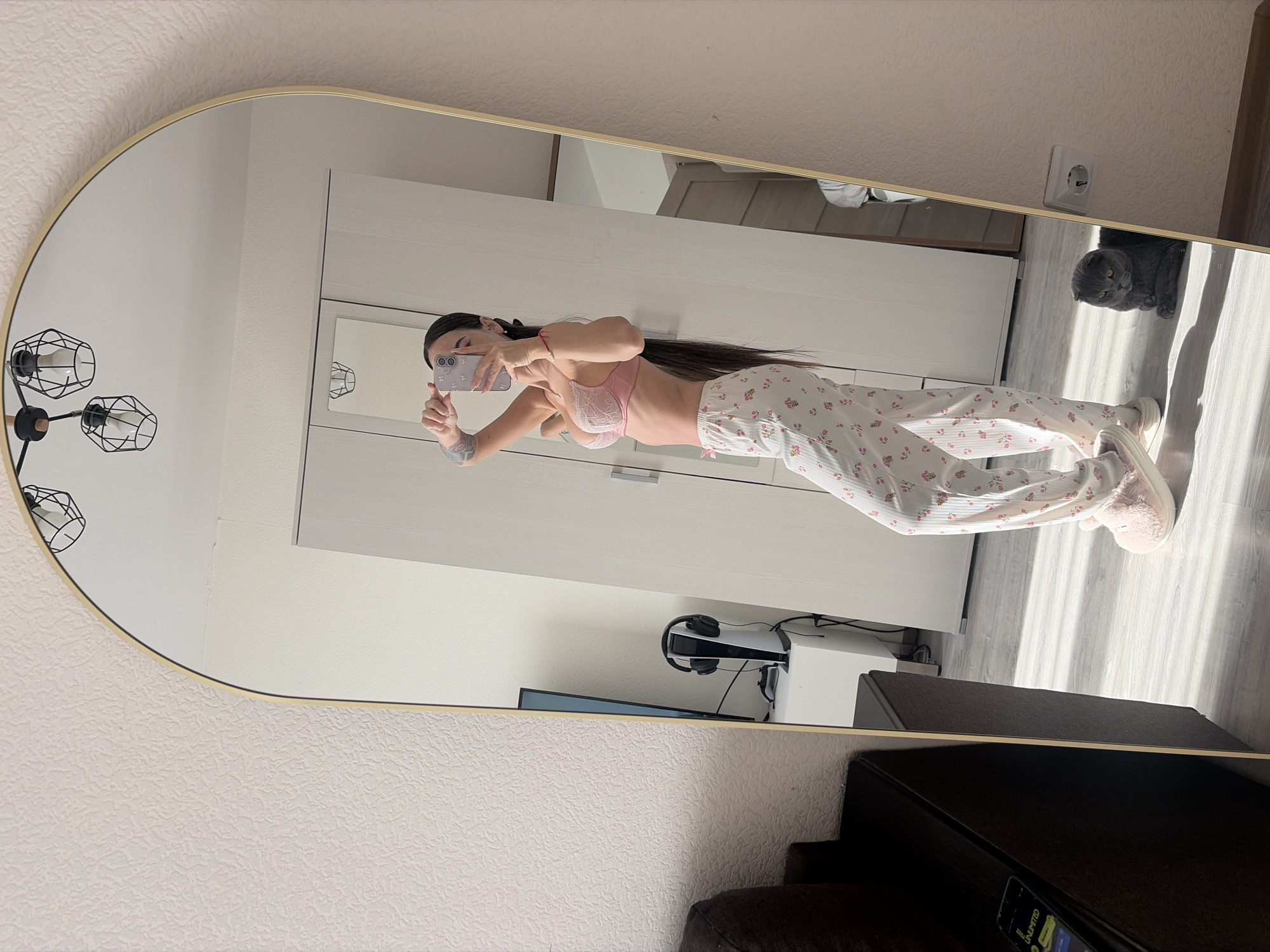 A young woman is seen in a mirror in what appears to be an indoor setting, possibly a private residence. She is holding a smartphone and taking a picture of herself. She is wearing a pink bra and patterned pajamas. The room contains furniture, including a wardrobe, a desk with a gaming console, and a chair. A cat is present in the reflection, lying on the floor. Based on the style of the interior decoration, particularly the light fixtures and the overall design aesthetic, the location could be Saint Petersburg, Russia.