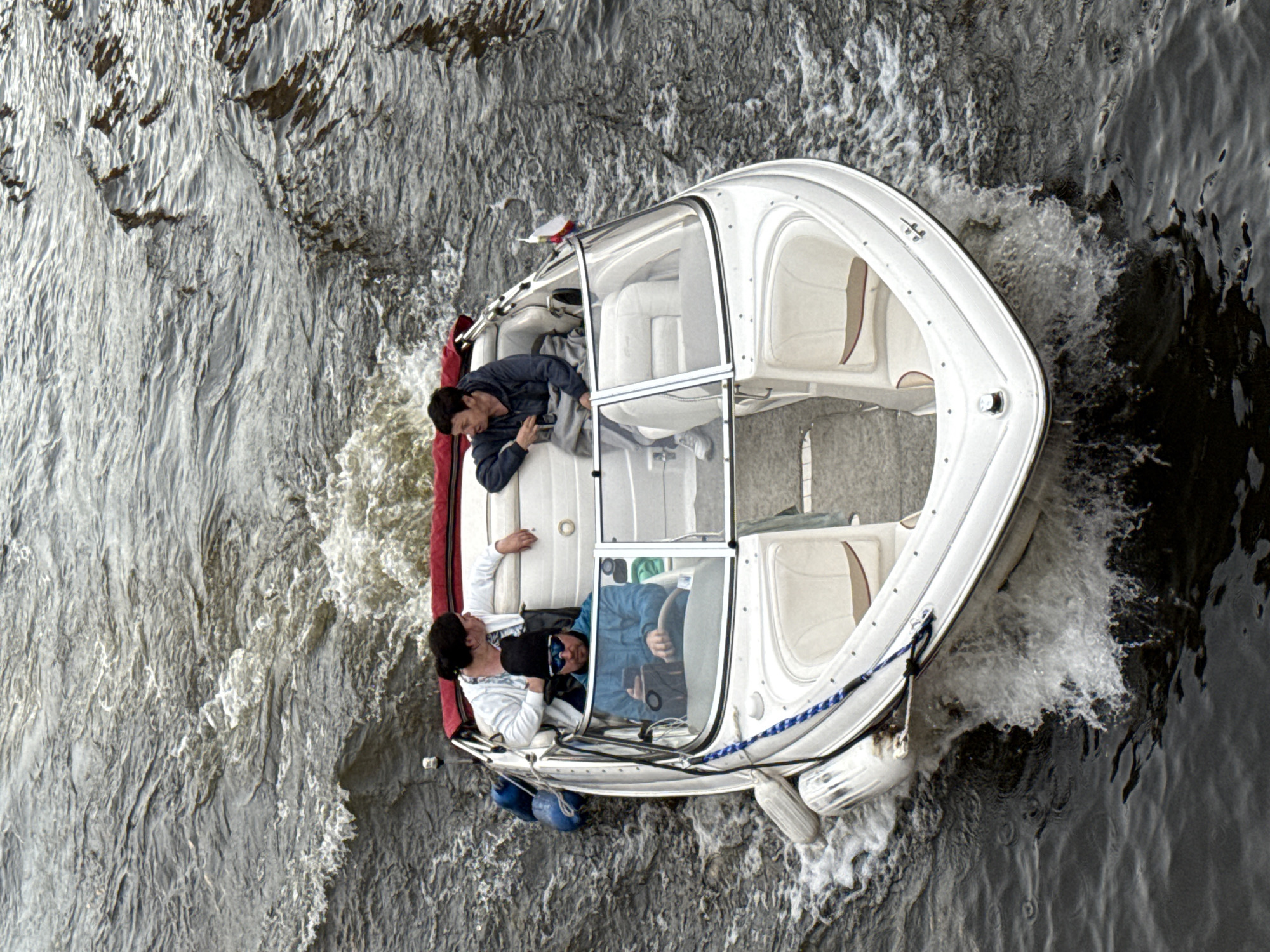 In this aerial shot, a white speedboat navigates through choppy, grey water, leaving a white wake. The boat, with its sleek design and white cushioned seating, appears to be moving at a moderate speed. Inside, three individuals are visible. One person, wearing a blue jacket, is seated at the helm, looking forward. Another person, in a grey hoodie, leans back in the passenger seat, holding a phone. A third individual, wearing a white hoodie, is reclining on the back of the boat. The scene suggests a leisurely outing on a waterway, possibly a river or a canal, given the location context of Saint Petersburg, Russia. The overcast sky and choppy water might indicate a cool, breezy day. The overall atmosphere seems relaxed and casual, with the individuals enjoying their time on the water. The water's surface is textured with ripples and splashes, hinting at movement and possibly some wind. No visible text is present in the image.