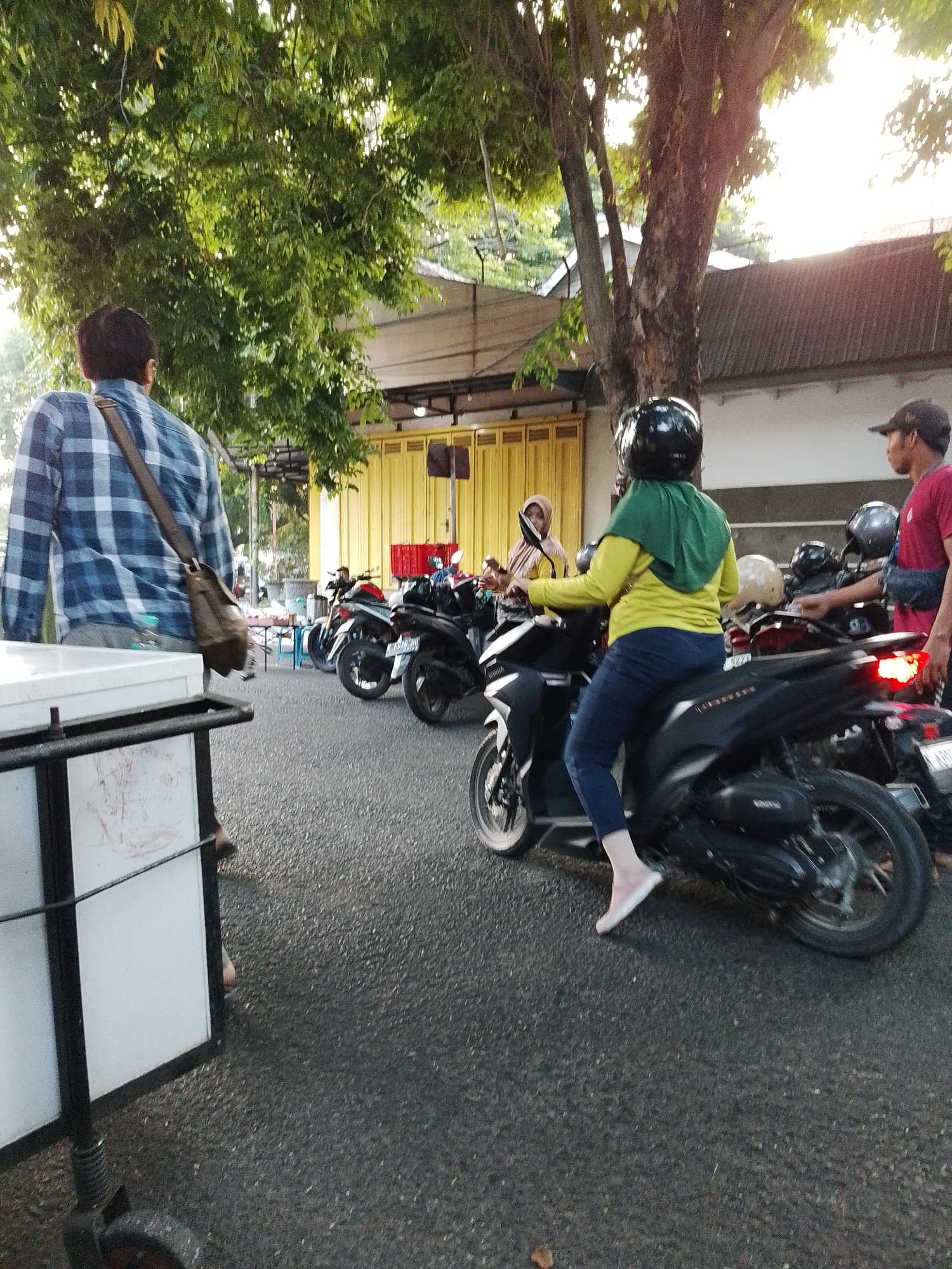 The image depicts a street scene in Kecamatan Pati, Indonesia, likely during daylight hours, possibly late afternoon due to the low angle of the sun. In the foreground, a person wearing a blue and white checkered shirt and a brown messenger bag is partially visible from the back, standing next to a white food cart. Further back, several motorcycles are parked on the asphalt road. One motorcycle is prominent in the mid-ground, ridden by a person wearing a green headscarf and a yellow jacket, with blue pants and white slip-on shoes. This rider is facing away from the camera, looking to their left. To the right of this motorcycle, another person wearing a red t-shirt and a dark baseball cap is standing and looking towards the motorcycles. In the background, a building with yellow doors is visible, and beyond that, trees and a roofline. The overall impression is a busy, outdoor environment, possibly a market or a roadside eatery.