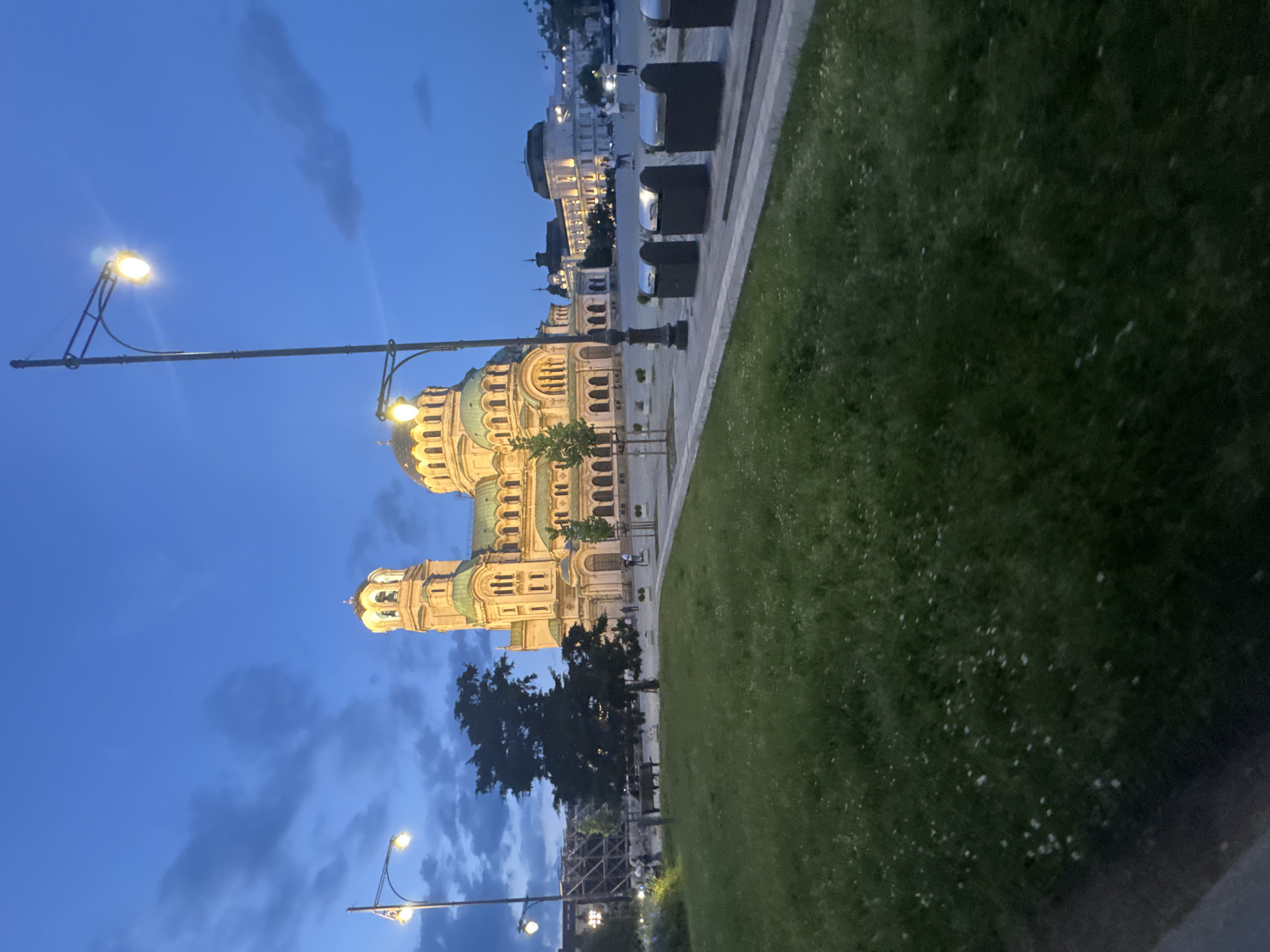 This image depicts the Alexander Nevsky Cathedral in Sofia, Bulgaria, at dusk or early evening. The sky is a deep blue with scattered grey clouds, and artificial illumination highlights the scene.
The cathedral, a prominent Neo-Byzantine structure, dominates the central and right portions of the image. Its multiple domes, including a large central dome and several smaller ones, are covered in a greenish material and are brightly illuminated with warm yellow light, as is the entire stone facade. A tall bell tower is visible on the cathedral's left side.
In the foreground, a large grassy area occupies the lower left quadrant, displaying numerous small white specks, possibly flowers. A paved walkway separates the grassy area from the street on the right. Along this paved area, a row of five dark grey waste bins with reflective silver tops is visible, extending towards the right.
Several streetlights with bright, yellowish-white lights are positioned throughout the scene. One tall streetlight stands prominently on the mid-left, and another is visible further right, closer to the cathedral.
A large, dark-leafed tree is situated in the mid-ground to the left of the cathedral, partially obscuring a park bench nearby. Smaller trees and shrubs are also visible around the cathedral and along the pathways.
In the background, beyond the cathedral and to its right, other multi-story urban buildings are illuminated. On the far left, a structure that appears to be scaffolding or a building under construction is partially visible.
Several people are dispersed throughout the scene. A small group is discernible near the tree and bench on the left, and other individuals are seen walking on the paved areas near the cathedral and the background buildings. No specific interactions among them are clearly visible.