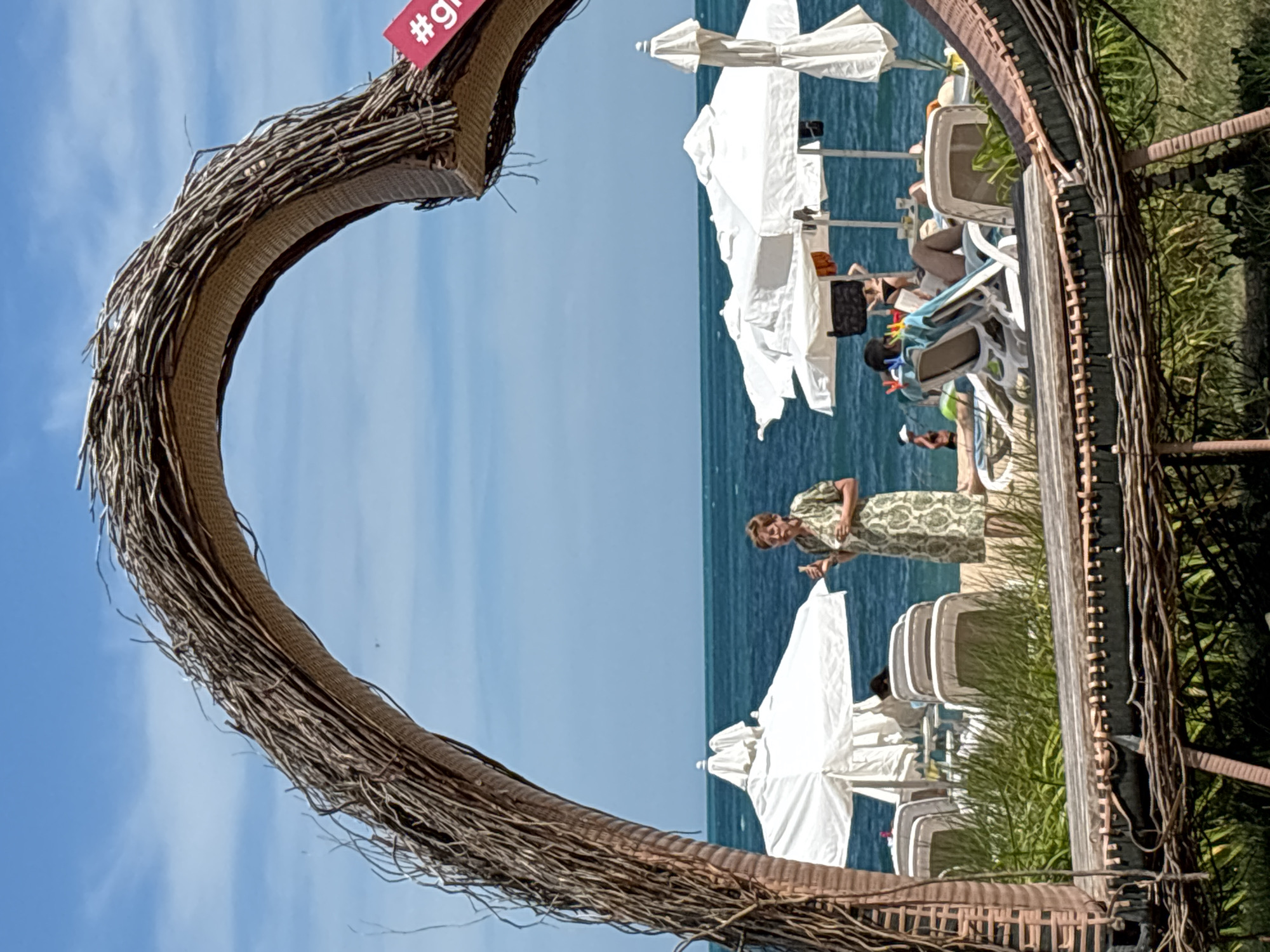 The image depicts a beach scene in Varna, Bulgaria, framed by a large heart-shaped structure made of woven branches. The sun is shining brightly, indicating it is daytime, with a clear blue sky and calm, blue ocean water. Several white beach umbrellas are set up, and some beach chairs are visible, with a few people relaxing on them. In the mid-ground, a woman in a patterned dress is standing and looking towards the viewer, while another person is partially visible behind her. The overall atmosphere appears to be one of relaxation and enjoyment on a sunny day at the beach. A small red tag with white text, "#gr", is attached to the heart-shaped structure.