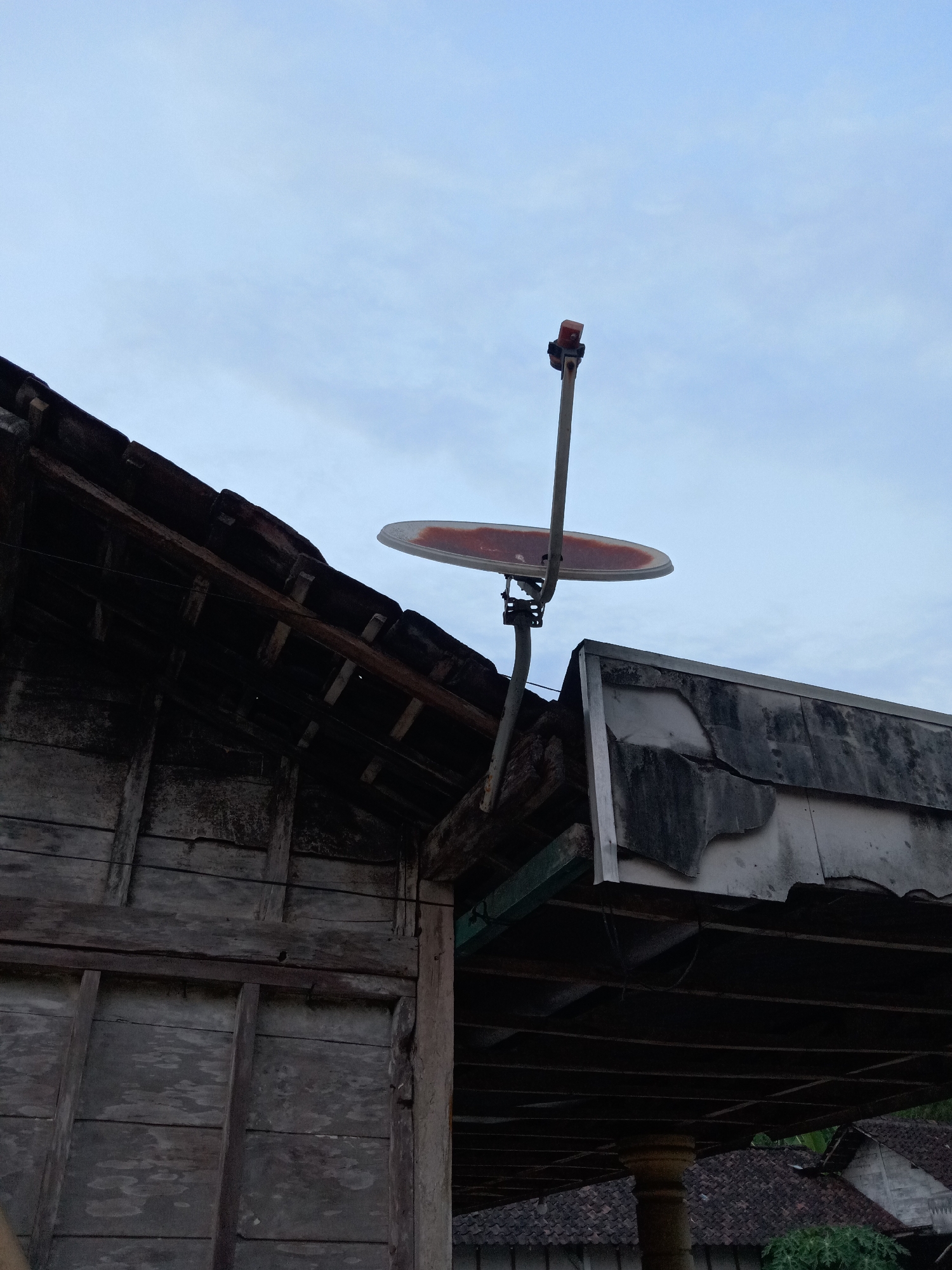 A satellite dish is mounted on a pole extending from the roof of a weathered, wooden structure. The dish is facing upwards and to the right, with a red component visible at its apex. Adjacent to the wooden structure is another building with a damaged, dark-colored roof. The sky is overcast with a light blue hue. The scene appears to be outdoors in a rural or semi-rural setting, possibly in Jumput, Indonesia, given the architectural style and vegetation that can be partially observed in the background.