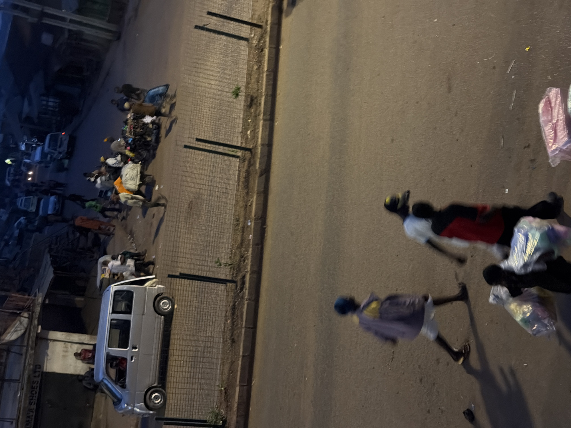 The image, taken in Bukuru, Nigeria, shows a street scene at night. The perspective is from above, looking down at the road and a paved sidewalk.
On the right side of the road, three people are visible. One individual in the foreground is wearing a purple shirt and white shorts, walking away from the camera. Behind them, two other individuals are lying on the ground, one appearing to be wearing a red and black shirt, and the other in white. Several plastic bags, some containing colorful items, are scattered near them. The lighting suggests it is nighttime, with artificial lights illuminating the scene.
On the left side of the road, a silver minivan is parked. Several people are gathered around the vehicle and along the sidewalk. There are also numerous objects, possibly goods for sale or discarded items, piled up on the sidewalk. In the background, there are buildings and more vehicles, contributing to a busy street environment. The overall atmosphere appears to be one of activity or perhaps a makeshift market at night. The visible text on a building reads "IMA SHOES LTD".