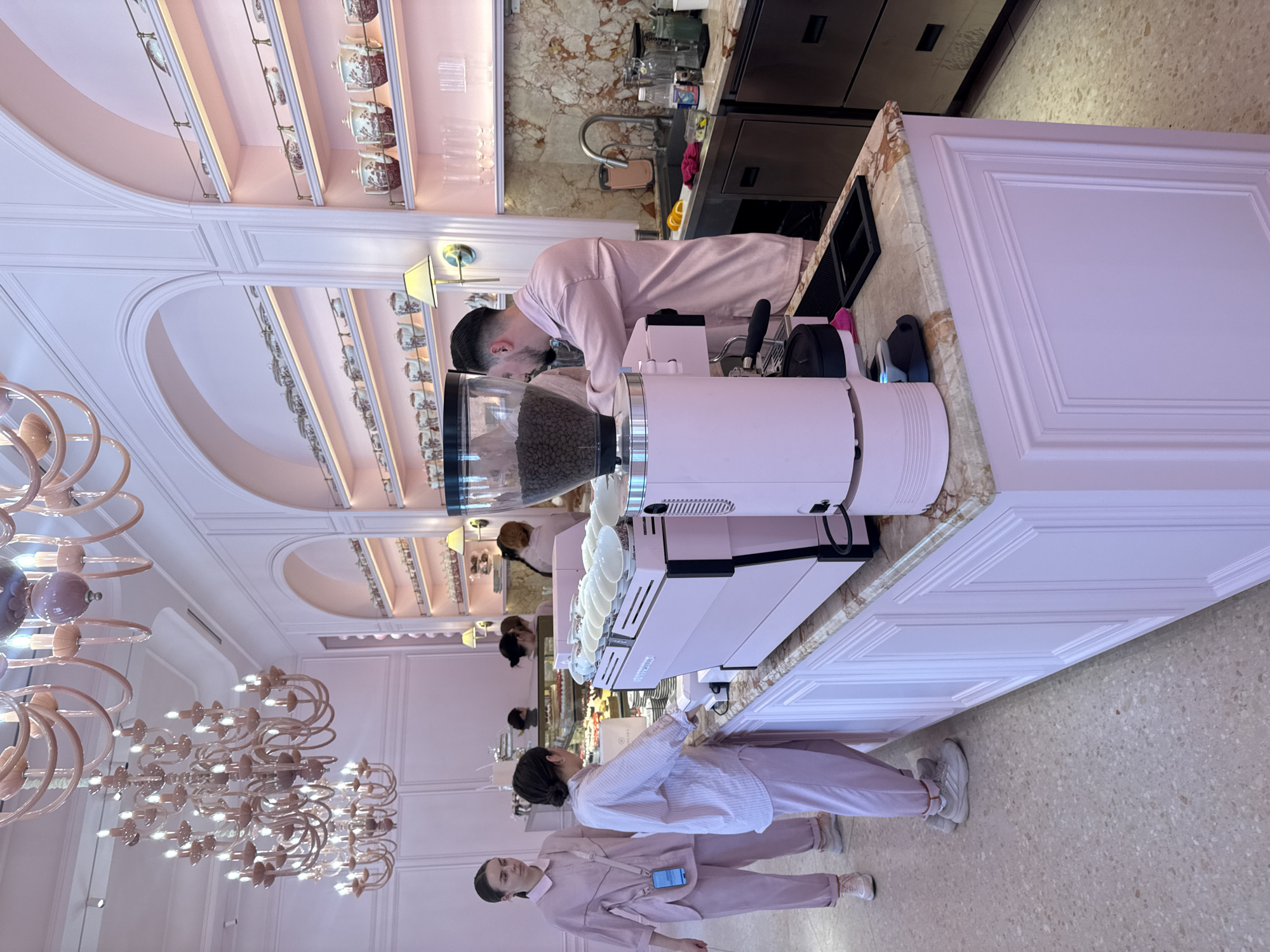The image depicts a brightly lit, pink-themed cafe interior, likely a pastry shop or dessert bar, located in Kyiv, Ukraine. In the foreground, a man with dark hair and a beard, wearing a pink button-up shirt, is operating a commercial-grade pink espresso machine. He is facing the machine, his hands are near the portafilter. To his left, two women, also dressed in pink, are standing at the counter. The woman on the left, with dark hair pulled back, is looking towards the right side of the frame. The woman on the right, with dark hair tied up, is looking at the counter and the espresso machine. She appears to be interacting with the barista. The counter itself is marble-topped and supported by pink cabinetry with decorative paneling. Behind the counter, shelves are lined with teacups, teapots, and other decorative items. The cafe features ornate chandeliers hanging from the ceiling. The overall aesthetic is elegant and feminine, with a prominent use of pink and gold accents.