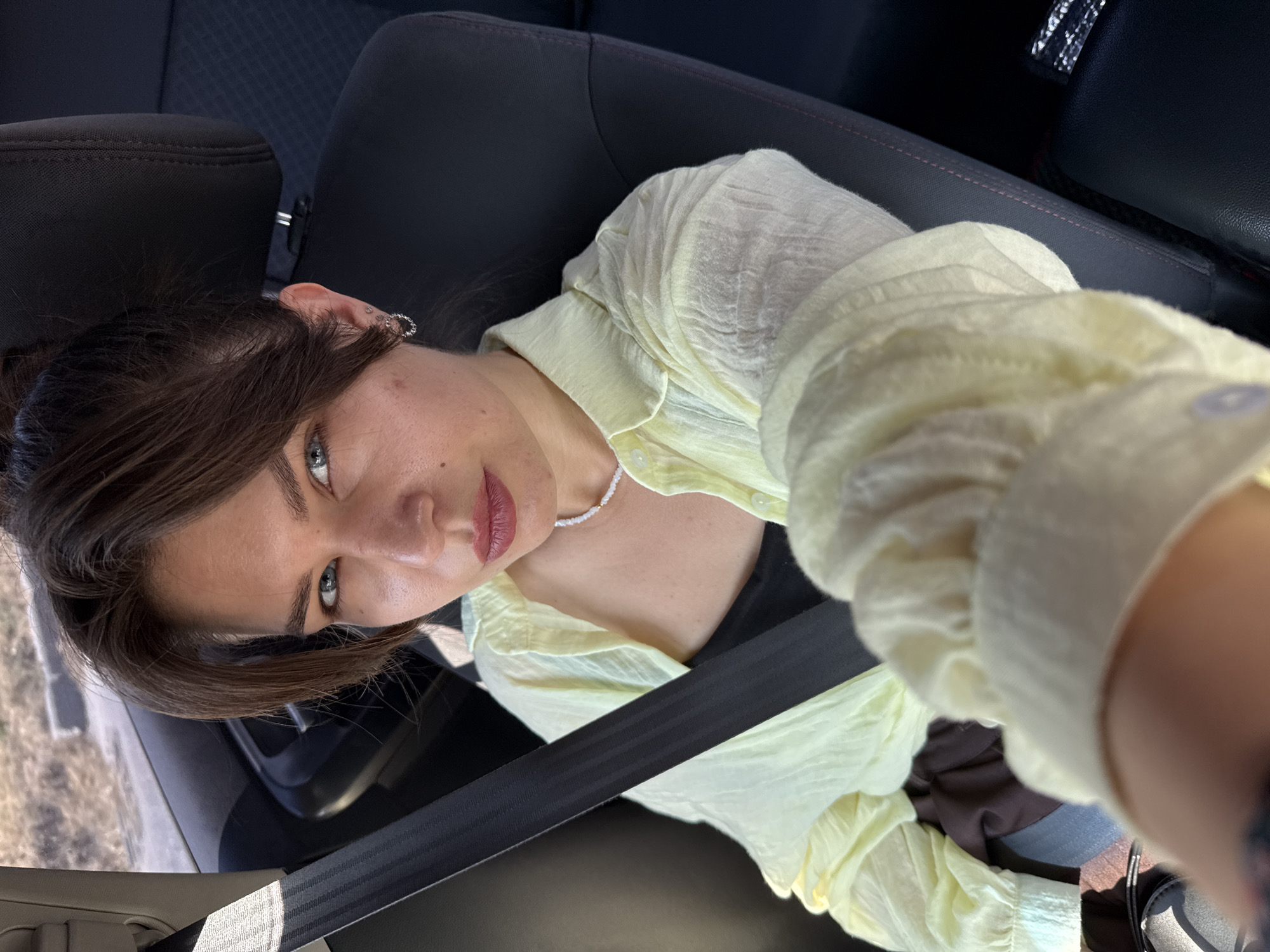 A woman is seen from a high angle, sitting in the passenger seat of a car. She has dark hair, styled loosely and falling around her face. She is wearing a light yellow, collared shirt with ruffled sleeves and a black top underneath. A delicate pearl necklace is visible around her neck. Her left ear is adorned with a hoop earring and a smaller stud. Her makeup includes subtle eyeliner and lip color. The seatbelt is fastened across her chest.
The setting appears to be daytime, with natural light illuminating the scene. The background shows the interior of a car, with dark upholstery and the edge of a headrest. Outside the car window, a blurry landscape is visible, suggesting the car is in motion. The overall tone of the image is casual and candid. There is no visible text in the image.