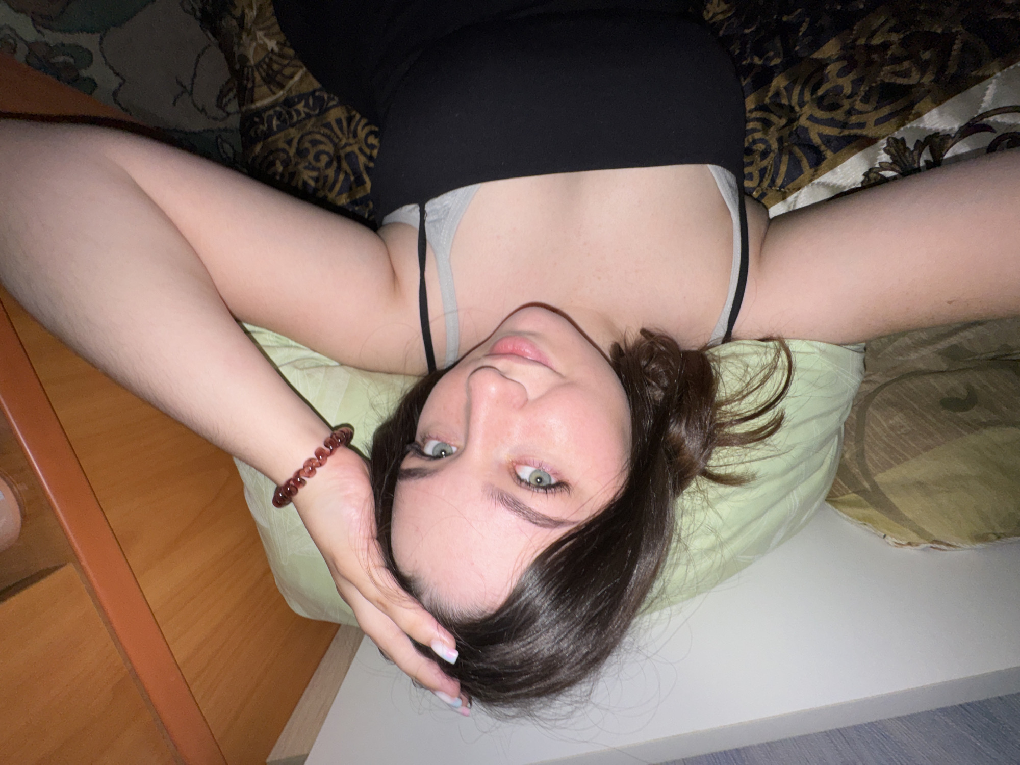 The image shows a young woman lying on her side on a bed. She has dark brown hair and blue-green eyes. She is wearing a black tank top and a grey bra. She is also wearing a red beaded bracelet on her left wrist. Her right arm is bent at the elbow, and her hand is resting on her head. The background is a bed with a light green pillow and a patterned comforter. A wooden surface is visible in the upper left corner of the image, possibly a nightstand or a shelf. The scene appears to be an indoor residential setting. The location of Бородулино, Russia is not depicted in the image.