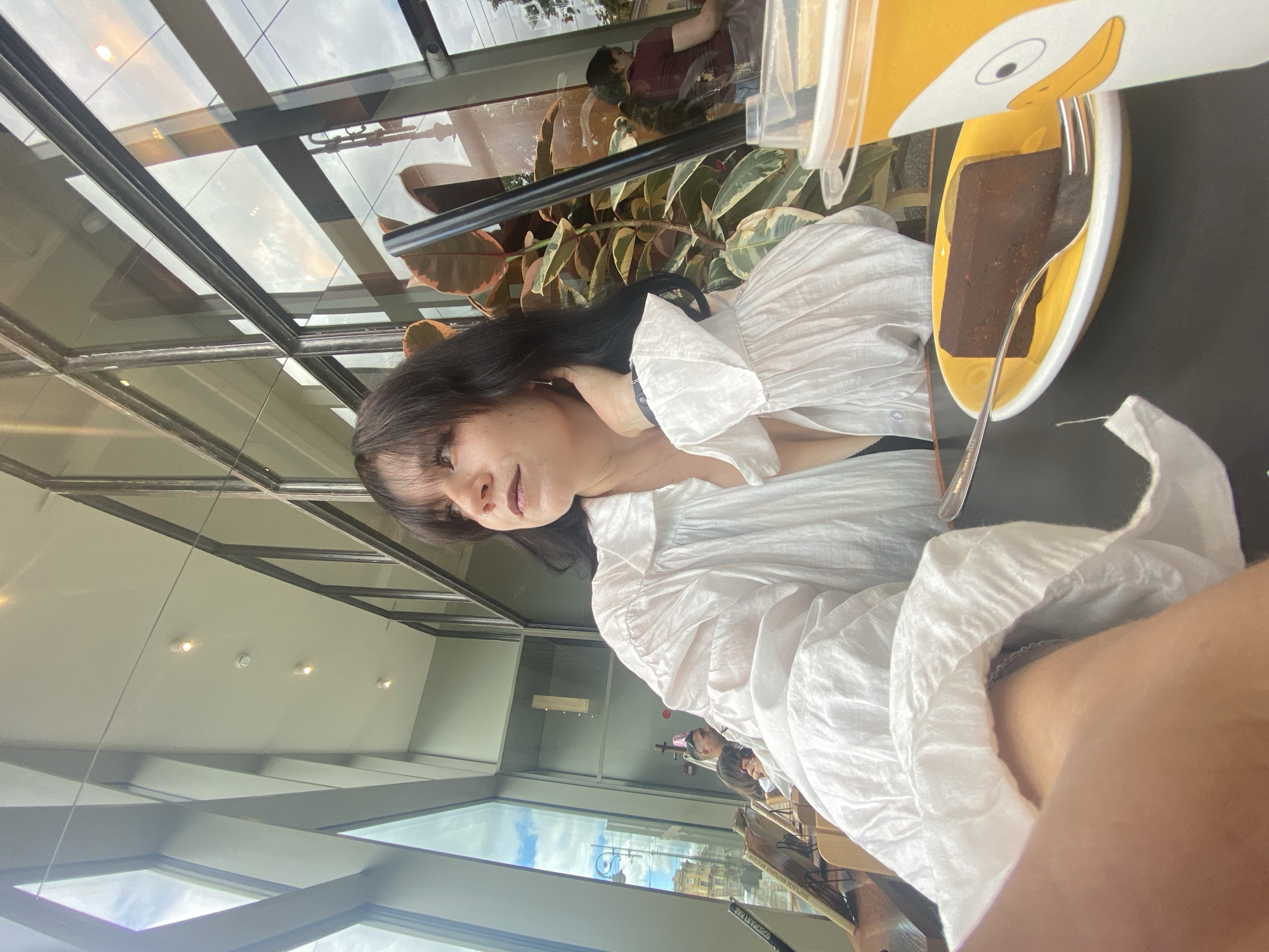 In Saint Petersburg, Russia, a young woman with dark hair and bangs sits at a table in a cafe. She is wearing a white, voluminous, ruffled shirt. In front of her, on a yellow plate, is a slice of chocolate cake with a fork. To the right, a cup with a cartoon duck design sits on the table. Behind her, a large ficus plant is visible, and through the large windows, the reflection of the sky and buildings can be seen. Other patrons are visible in the background, some seated and some standing.