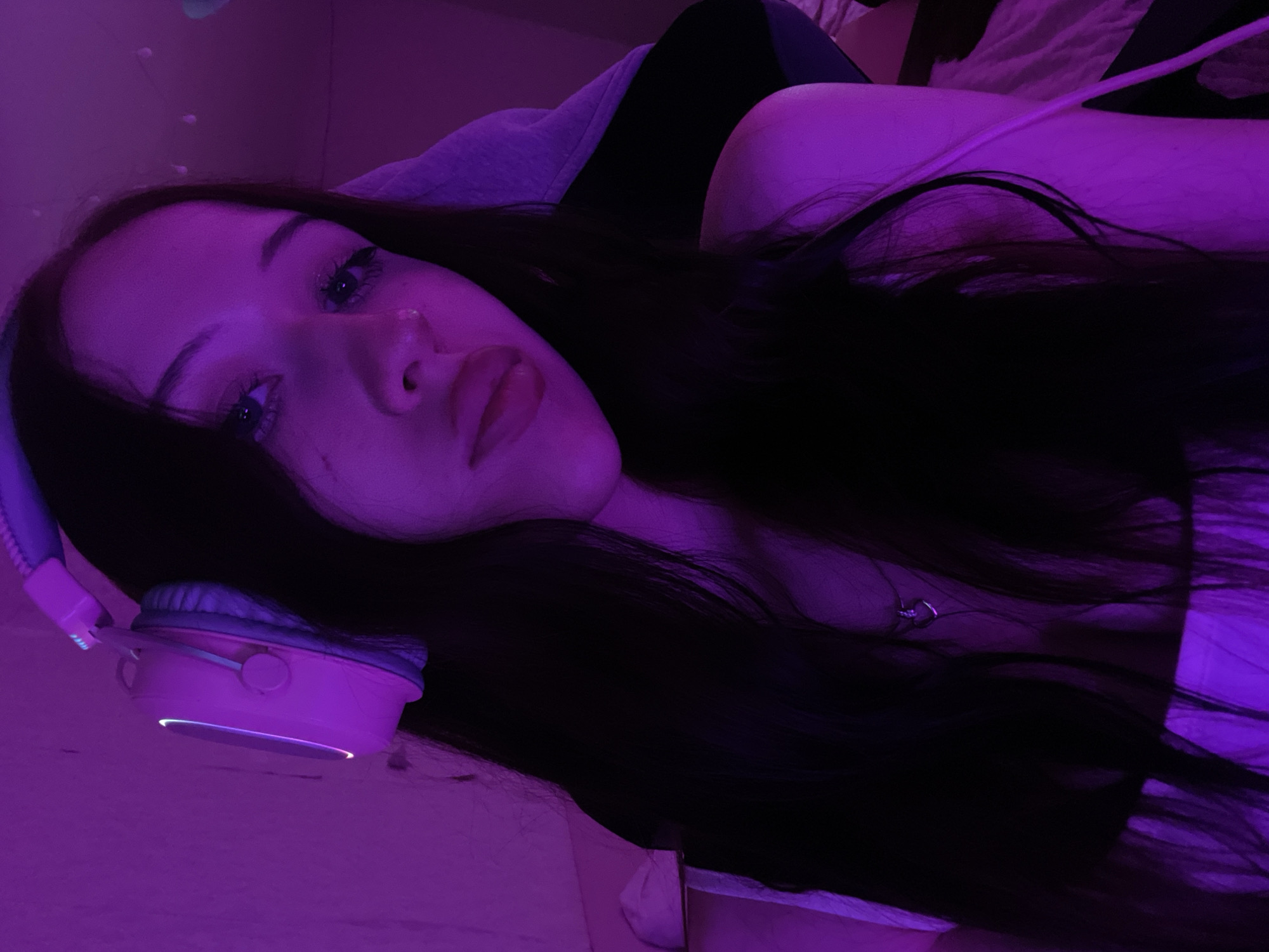 The image depicts a young woman with long, dark hair wearing pink headphones. She is illuminated by purple lighting, which casts a dim glow on her face and surroundings. She appears to be indoors, possibly in a room with a textured wall visible in the background. The lighting suggests a low-light environment, common in gaming setups or studios. No specific location in Votkinsk, Russia, can be identified from the image.