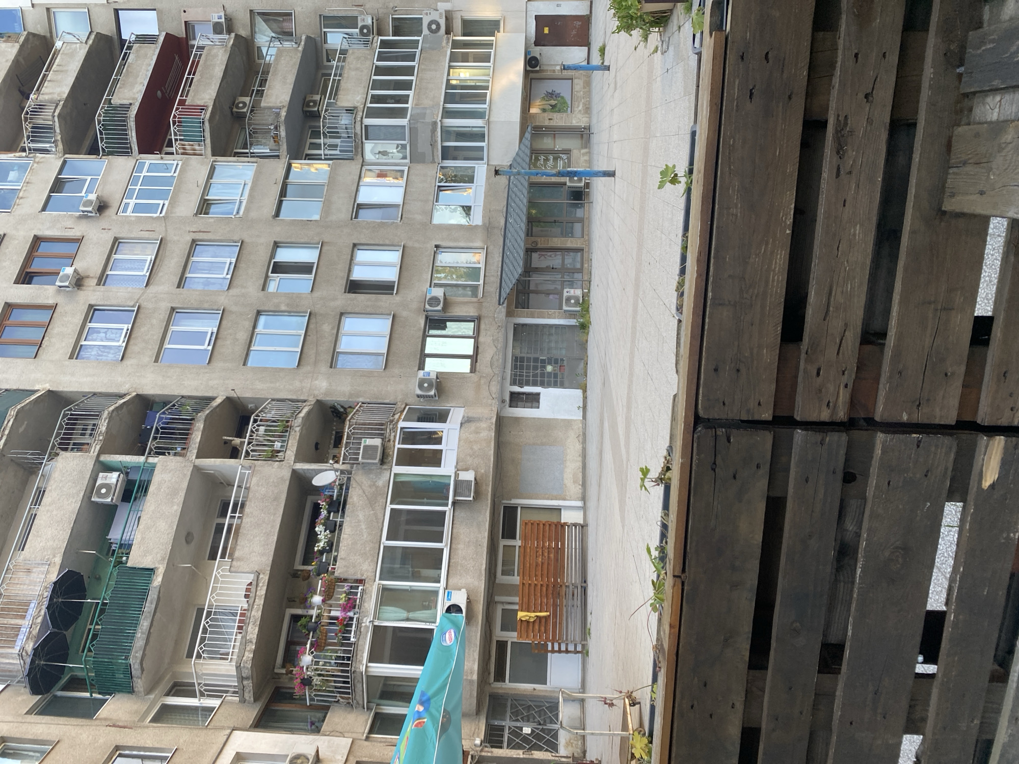 The image shows an exterior view of a building and adjacent objects in Sofia, Bulgaria. The building appears to be a multi-story apartment complex with numerous windows and balconies. Air conditioning units are visible on many of the balconies and near windows. The facade is made of concrete and painted a light gray color.
On the right side of the image, wooden pallets are stacked vertically. In the foreground, the lower part of a Nestlé-branded umbrella can be seen. Further back, a small seating area with a brown bench is located near one of the ground-level entrances. There is a sidewalk in front of the building. The image is rotated counter-clockwise.