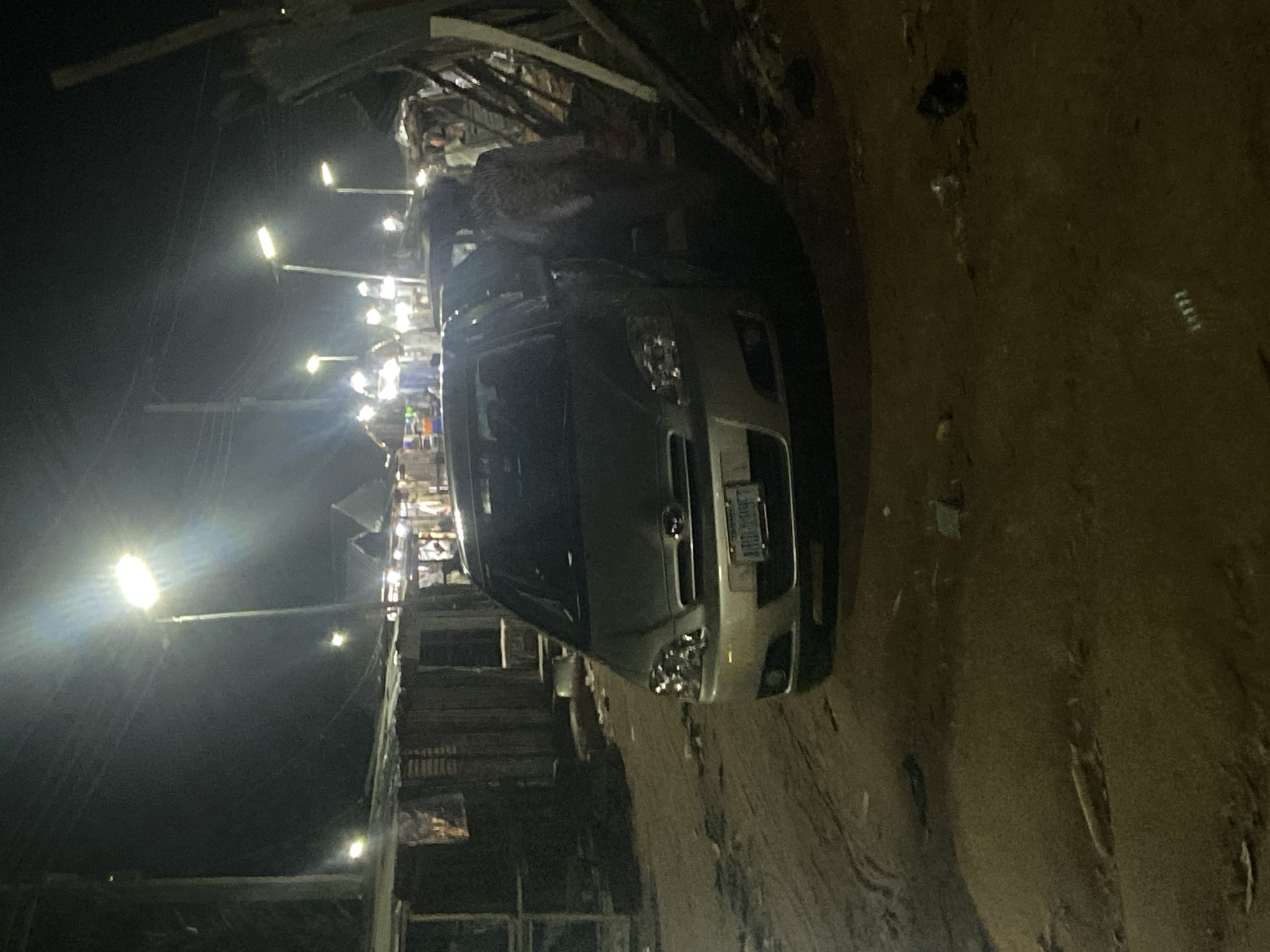 The image captures a scene at night in Kuson, Nigeria, likely a market or a busy street with various structures. The primary subject is a light-colored car parked on a dirt road. The car's headlights are illuminated, and its front grille and license plate are visible. Behind the car, a cluster of stalls or buildings is illuminated by streetlights, creating a vibrant, nighttime atmosphere. Several tall streetlights are visible, casting a bright glow on the surroundings. The background shows more structures, possibly market stalls or shops, with some people faintly visible in the distance, engaged in activities within the illuminated areas. The ground appears to be unpaved, indicating a rural or semi-urban setting. The overall impression is of a lively night scene in a community, possibly a local market that operates after dark. The image is a bit tilted, with the car appearing to be on its side.