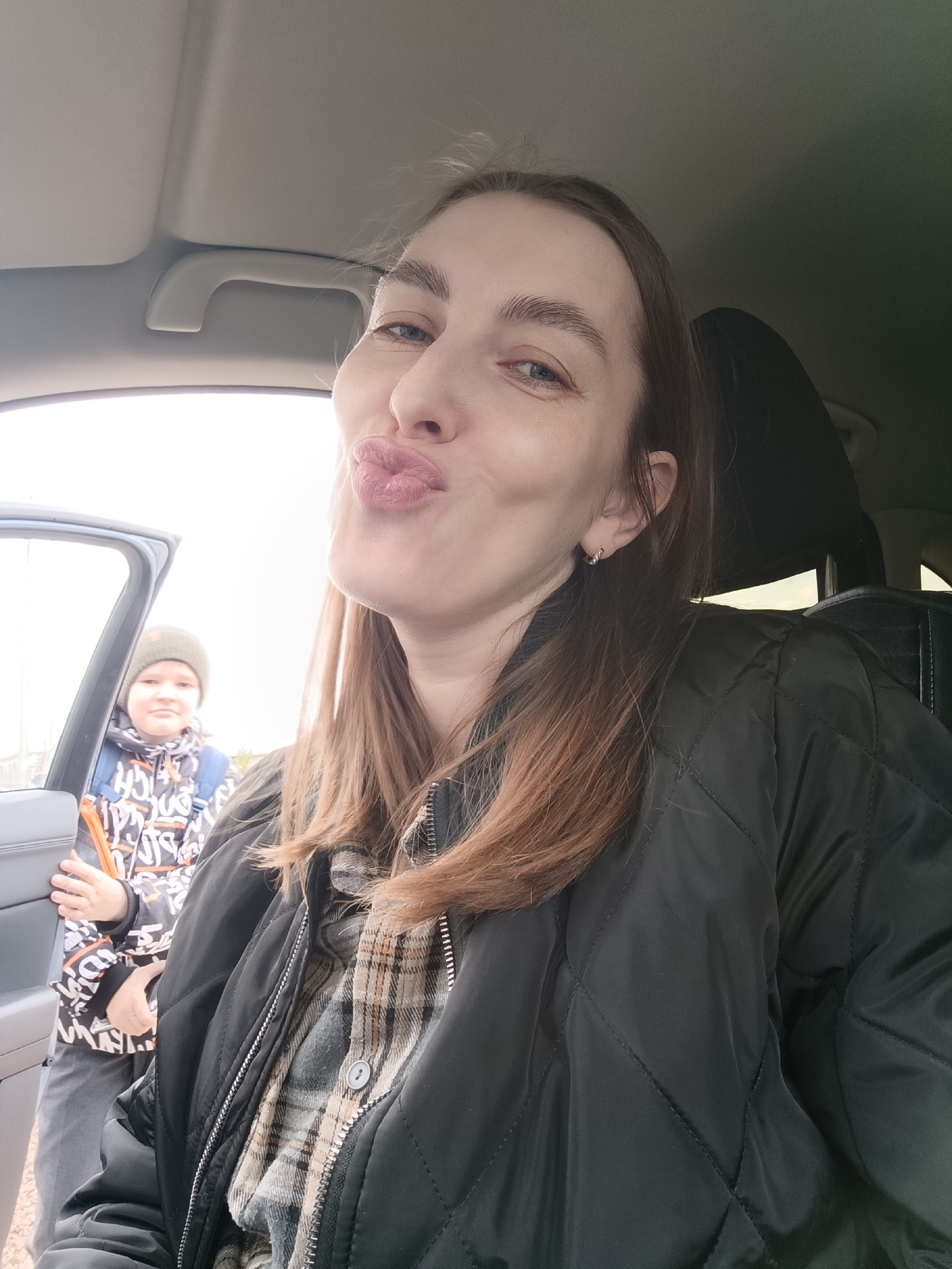 A selfie taken inside a car in Chaikovsky, Russia, features a woman in the foreground with a child visible by an open car door in the background. The woman, appearing to be in her late 20s or early 30s, occupies the majority of the frame, looking directly at the camera with her lips puckered in a playful "kissing" expression. She has long, straight light brown hair and light blue eyes, and wears a dark, possibly quilted, jacket over a plaid shirt. A small hoop earring is visible in her right ear.
In the mid-ground, partially framed by the open car door, a child, likely a boy, stands outdoors. He wears a grey beanie and a dark jacket adorned with white abstract patterns and some indistinct black letters or symbols, appearing to include "CH" and "G". A blue backpack is on his shoulders, and he holds a small orange object in his right hand. The child looks towards the camera with a neutral to slightly smiling expression.
The setting is daytime, with an overcast sky indicated by the bright, diffused light coming from outside the car. The car's interior, including the ceiling and a grab handle, is visible in the upper left. The jackets worn by both subjects suggest cool weather. The scene conveys a casual and familial moment.