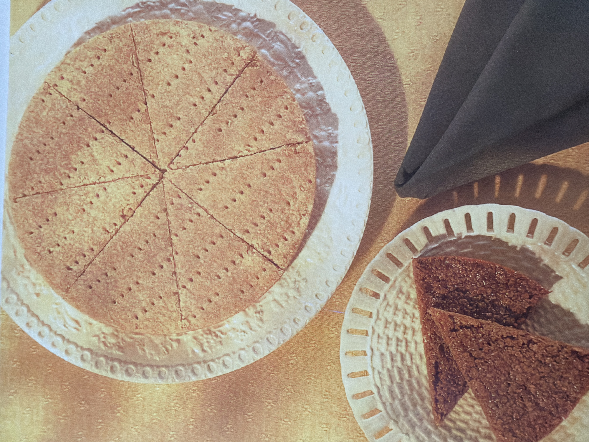This is a medium shot, eye-level, indoor still life photograph of two plates of baked goods on a textured, golden-brown surface. The plate on the left holds two triangular-shaped, dark brown cookies with a coarse texture. The plate on the right holds a circular, light brown baked item that has been cut into eight triangular wedges. The wedges are covered in small, uniform indentations. The plates are white with decorative, perforated edges. To the right of the plates, a dark navy blue napkin is folded diagonally. The lighting casts soft shadows on the surface. The scene is presented without any people or specific environmental context that would indicate a city or location.