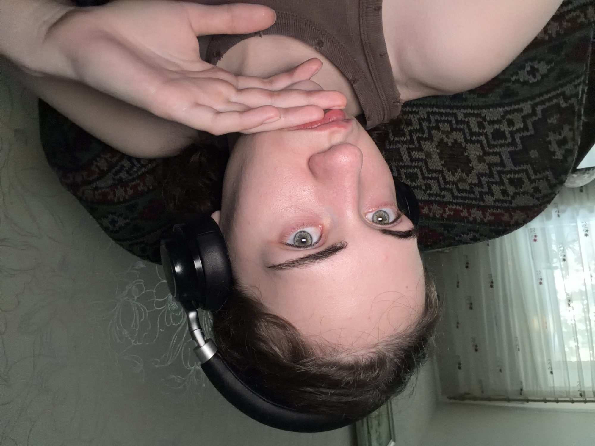 The media file shows a person lying down, with their head near the bottom of the frame and their feet near the top, due to the upside-down orientation of the image. The person is wearing a brown tank top and black headphones. Their right hand is raised to their face, covering their mouth and nose, and their eyes are open and looking directly at the camera. The person has light-colored eyes and dark eyebrows. The background appears to be indoors, with a patterned blanket or fabric on which the person is lying. To the right, a sheer white curtain with a floral pattern is visible, suggesting a window. The lighting is dim, indicating it might be evening or a dimly lit room. The person's expression is difficult to discern due to their hand covering their mouth, but their eyes convey a sense of directness or perhaps surprise. There is no visible text in the image. The location context is Zavodoukovsk, Russia.