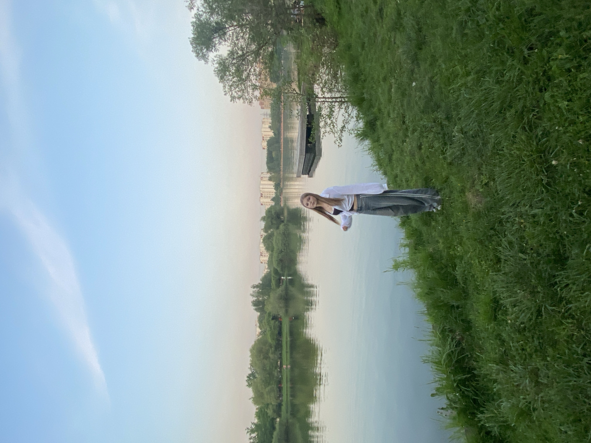 A young woman stands on a grassy bank beside a body of water, likely a lake or river, on an overcast day. She has long blonde hair, wears a white top and darker pants, and is angled slightly towards the camera. In the background, there are buildings and trees, suggesting a park or recreational area within a city. The buildings indicate a location in Moscow, Russia. A pier is visible over the water.