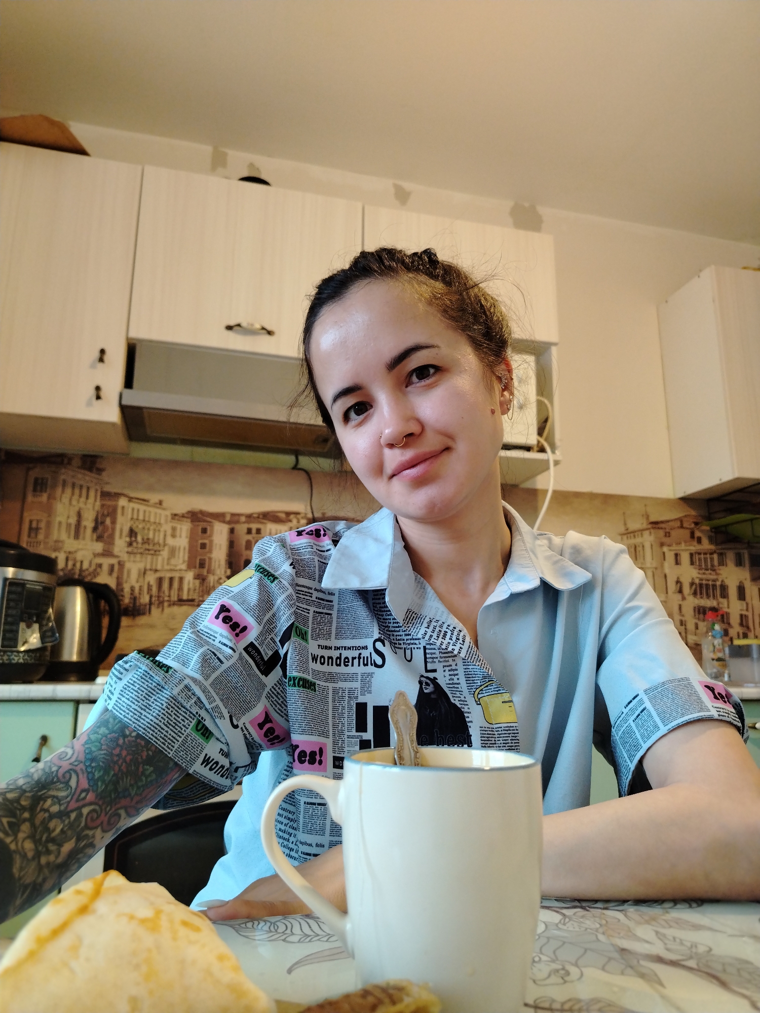A young woman sits at a kitchen table in what appears to be an apartment. She is fair-skinned with brown hair pulled back, a nose ring, and is wearing a light blue shirt with newspaper-printed sleeves. A white coffee mug sits in front of her, with a spoon inside. A plate of food, possibly pancakes, is also on the table. The kitchen has light-colored cabinets and a patterned backsplash. An electric kettle is visible in the background. The location cannot be verified as Noviy, Russia.