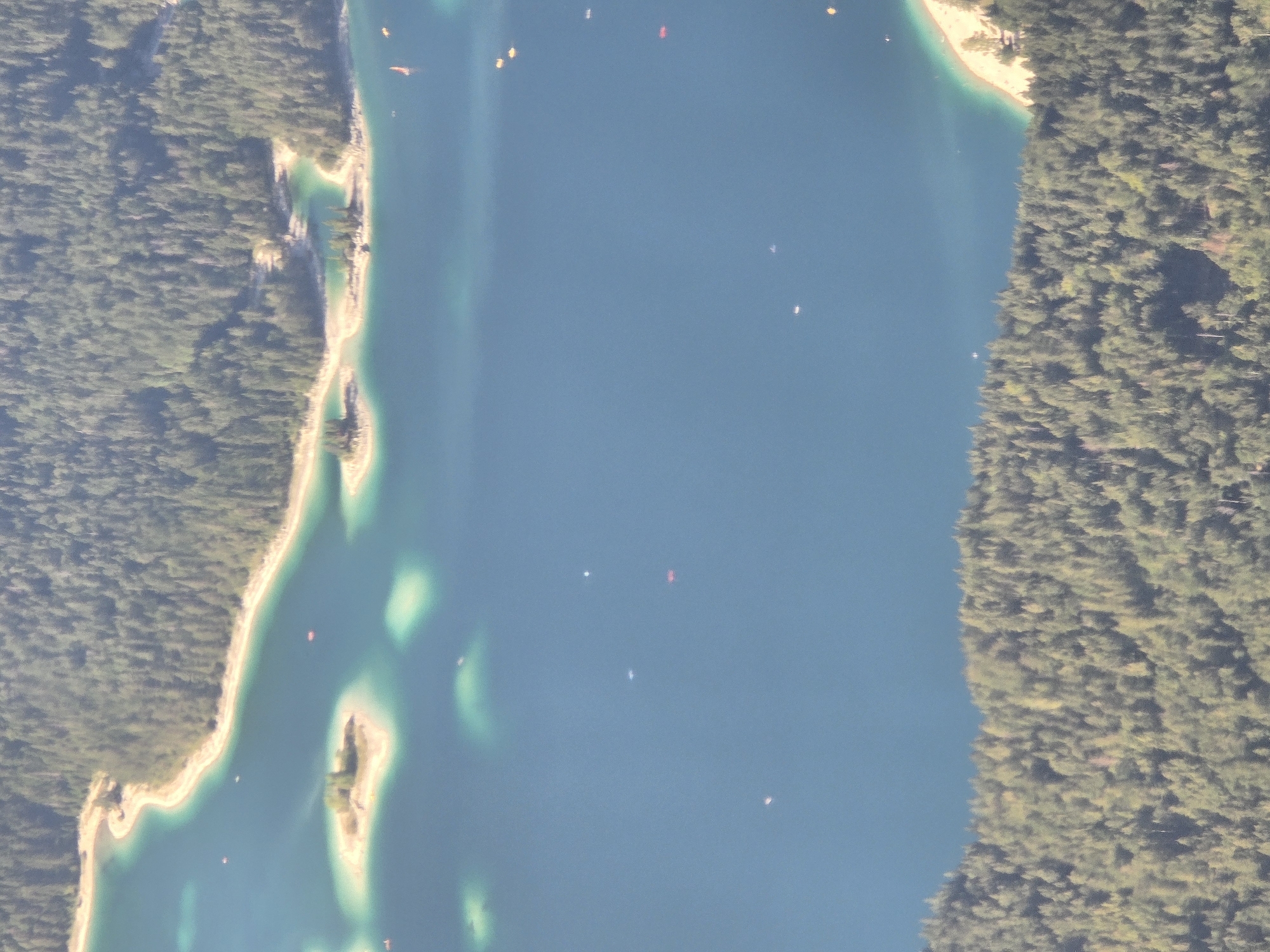 The image displays an aerial view of a clear blue lake, likely Eibsee, situated in Garmisch-Partenkirchen, Germany, surrounded by a dense, dark green forest. Several small, sandy islands with clusters of trees dot the lake's surface. The shoreline of the lake features a light-colored beach that transitions into the forest. The water appears calm, with a few small, indistinct objects that could be boats or buoys scattered across its expanse. The overall scene is one of natural beauty, with the vibrant blue of the water contrasting with the deep green of the trees.