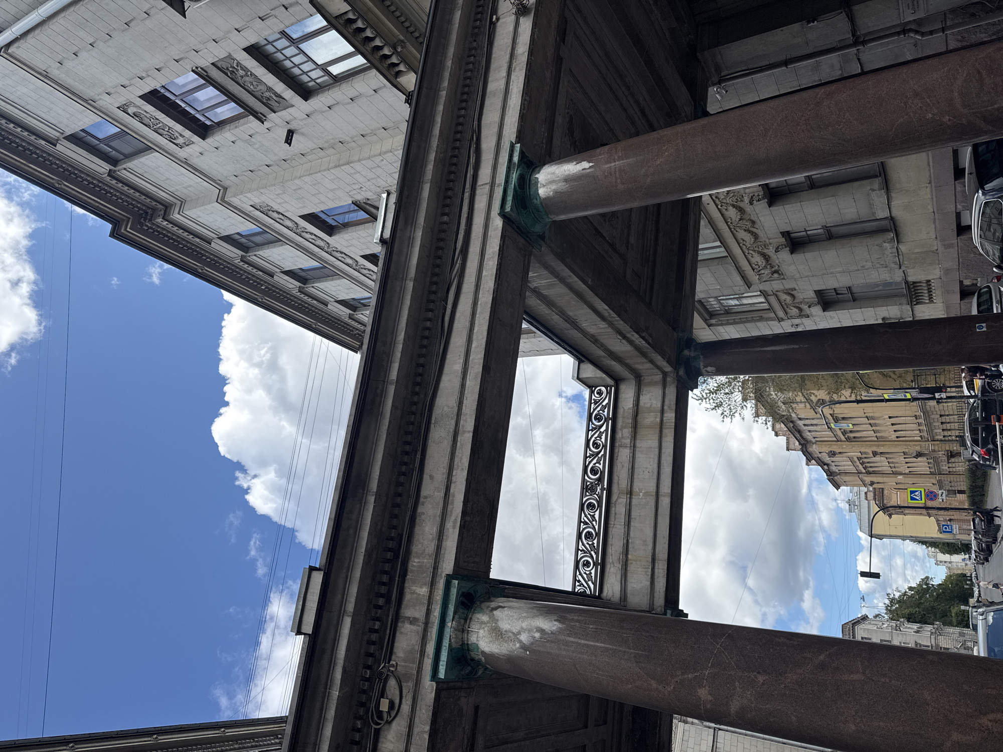 The image shows an outdoor, eye-level, slightly low-angle, full shot of an architectural structure with large marble columns supporting an overhead walkway. The walkway is adorned with an ornate iron railing. Beyond the structure, a street scene is visible with buildings lining both sides, parked cars, streetlights, and traffic signals. The sky above is partly cloudy with a bright blue sky.
This is a view of the arcades of the General Staff Building on Palace Square in Saint Petersburg, Russia. The General Staff Building is a monumental neoclassical building that flanks Palace Square along with the Winter Palace.