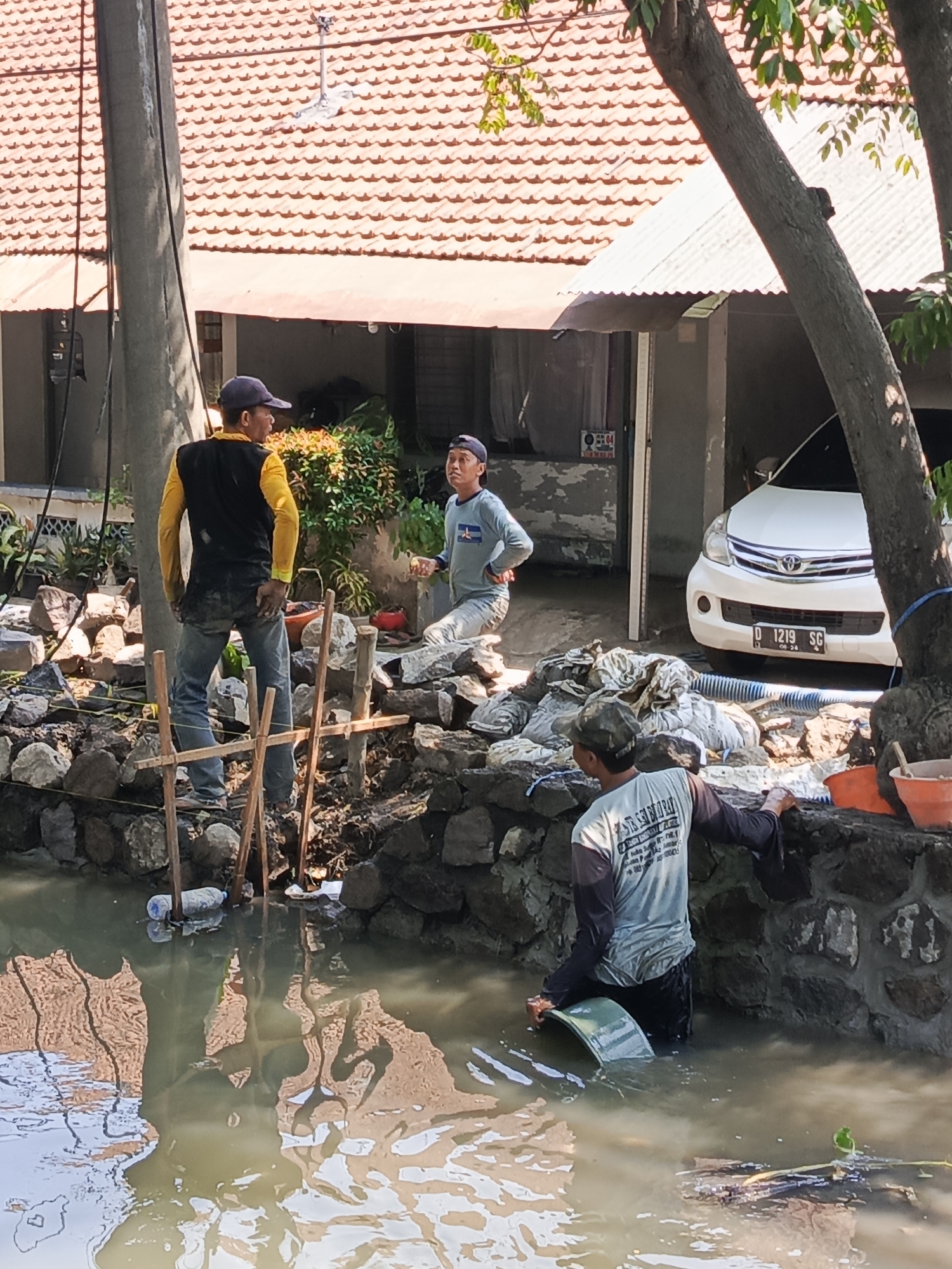 The image depicts a construction scene near a residential property in Pulungan, Indonesia. Three men are engaged in what appears to be canal or drainage work. In the foreground, one man stands in murky water, holding a curved object, possibly a tool or part of the construction material. Behind him, a stone wall is being constructed. Another man, wearing a yellow long-sleeved shirt and black vest, stands on the stone embankment, observing the work. To his left, a third man, in a light blue long-sleeved shirt, sits on the embankment, also looking towards the construction. A white car is parked in the background, with a house and tiled roof visible behind the construction area. The environment is sunny, with trees providing some shade. The water reflects the surrounding structures and sky.
