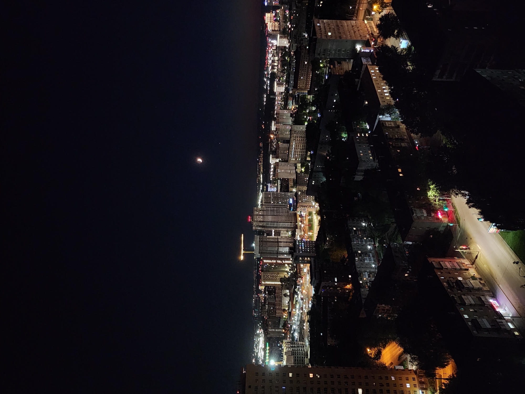 A nighttime, elevated view of a city. The left of the image is dominated by a dark expanse, likely the sky or a body of water, with a sliver of moon visible. The right side showcases the city itself, illuminated by numerous lights from buildings and streets. There are rows of buildings of varying heights, some with visible windows and street lights between buildings and possibly cars on streets.