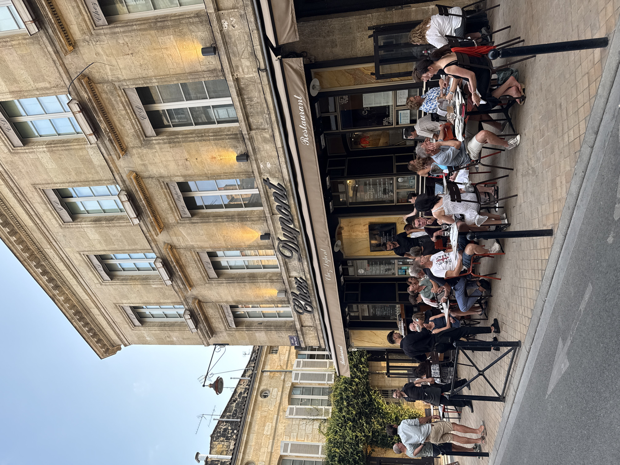 The image depicts an exterior view of the restaurant "Chez Dupont" located on a street in Bordeaux, France. The restaurant occupies the ground floor of a historic stone building. Several patrons are seated at tables on the outdoor patio, engaged in dining and conversation. Two servers, dressed in black, are also visible, attending to the customers. The street in the foreground is paved, and a metal fence separates the sidewalk from the road. The architectural style of the building, with its stone facade and windows, is characteristic of Bordeaux. The lighting suggests it is late afternoon or early evening.