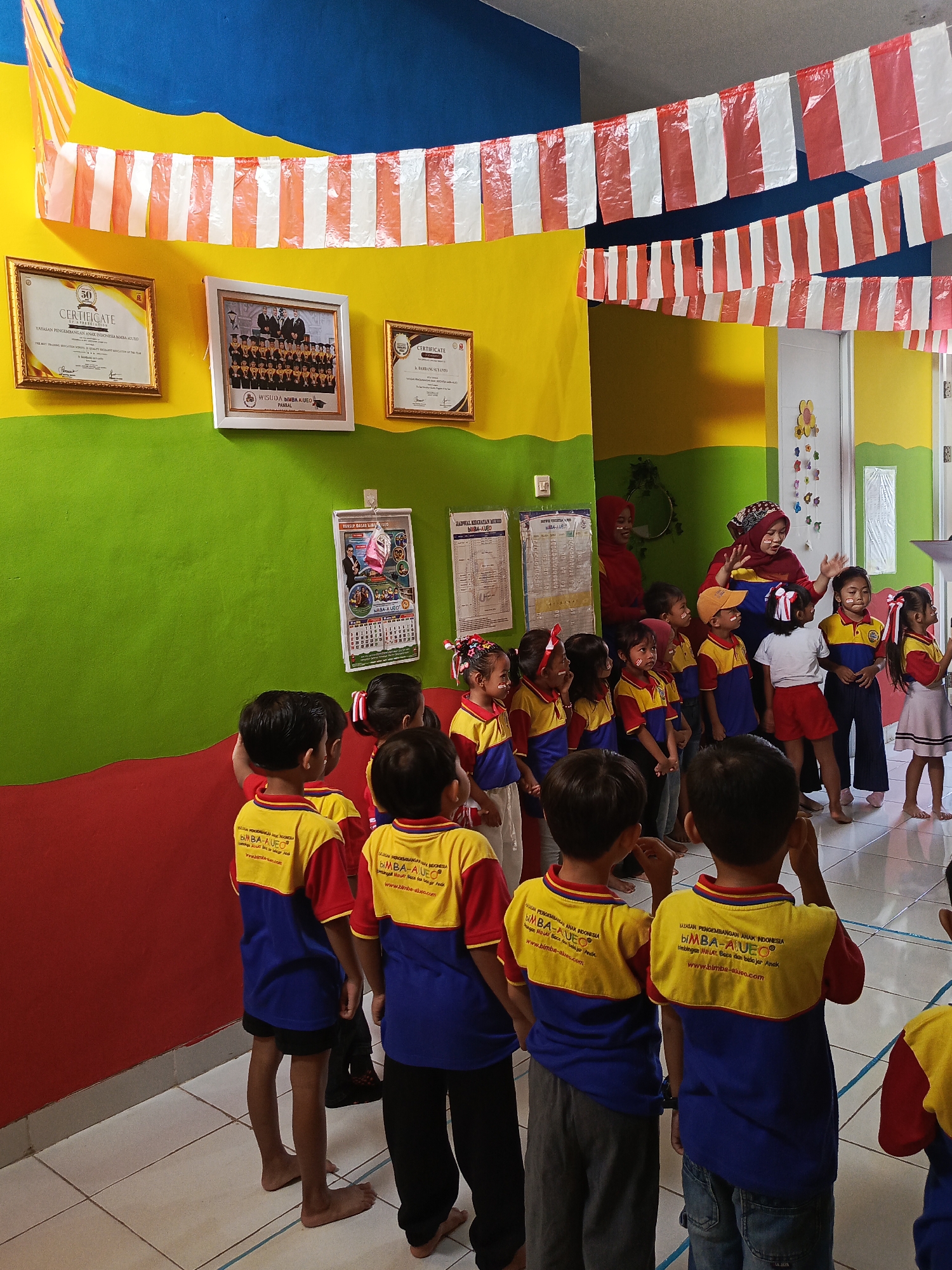 In Ciseeng, Indonesia, a group of young children, likely attending kindergarten, are gathered indoors. They are dressed in matching blue and yellow uniforms with red accents. Some of the children have face paint on their cheeks, suggesting a festive occasion. Two women, one wearing a red hijab, are also present, appearing to instruct or engage with the children. The room is decorated with red and white striped banners hanging from the ceiling and several framed certificates and a photograph are displayed on the walls. The walls themselves are painted in vibrant sections of blue, yellow, and green. The floor is tiled. The children are facing towards the two women, some with hands raised, as if participating in an activity or song. The overall atmosphere appears to be educational and celebratory.