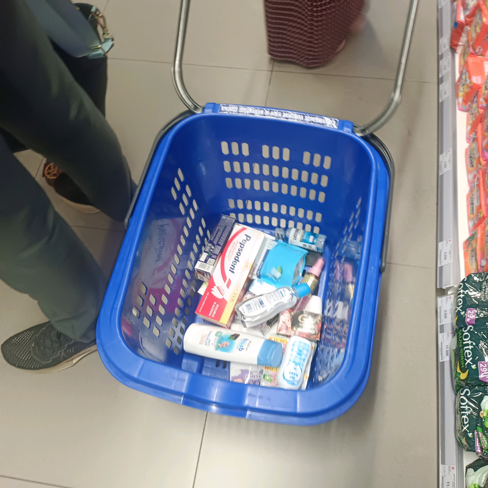 In the city of Surabaya, Indonesia, a high-angle, eye-level shot captures a blue plastic shopping basket filled with various personal care items, including toothpaste, soap, and other toiletries. The basket sits on a light-colored, tiled floor, with the legs of an unseen person visible in the lower left corner, wearing dark pants and black sneakers. A red and white striped bag is partially visible behind the basket, and to the right, a shelf is stocked with packaged sanitary products, including brands like "Softex." The lighting suggests an indoor retail environment, likely a supermarket or pharmacy.