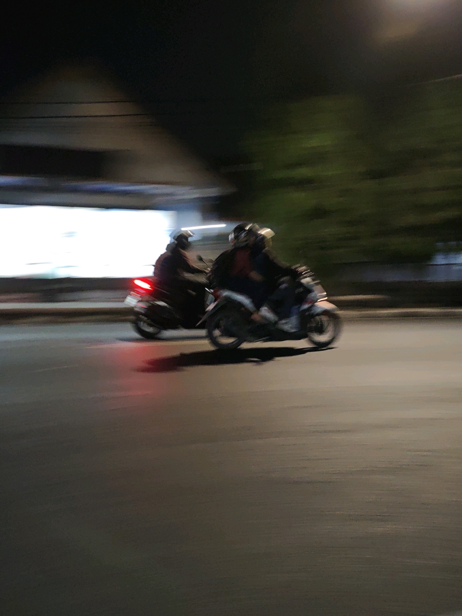 Two people on a scooter are captured in motion at night on a street in Kediri City, Indonesia. The scooter is dark-colored and the rear taillight is illuminated with a red glow, indicating it is in use. The riders are wearing helmets and appear to be traveling together. The background is blurred with dark foliage and a brightly lit rectangular structure, possibly a building or billboard, on the left side. The street itself is asphalt and reflects the ambient light. The image conveys a sense of movement and urban nighttime activity.