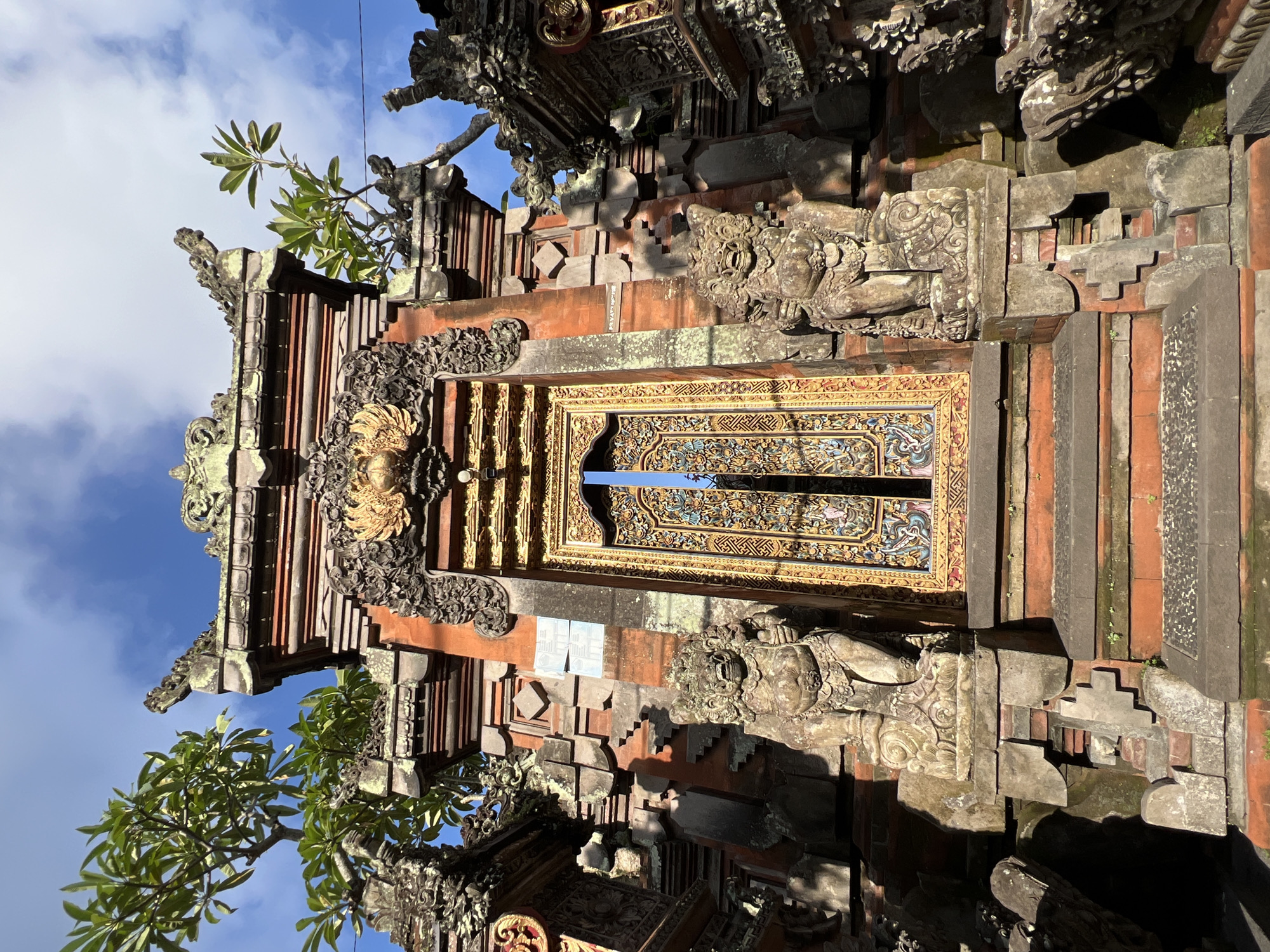A low-angle, close-up shot captures a richly decorated Balinese temple gate within the Ubud District of Indonesia. The gate features a grand wooden door, intricately carved and painted with gold, blue, and other vibrant colors, prominently displayed at its center. A blue sash or ribbon hangs from the upper part of the door.
Flanking the door are detailed stone guardian figures, possibly mythological creatures, their forms weathered but still distinct, with moss or lichen growing on parts of the stone. The temple structure itself is built from a combination of reddish brick or stone and grey, aged stone blocks, featuring multiple tiers and an abundance of elaborate carvings, reliefs, and sculptural details, typical of traditional Balinese architecture. Above the door and along the gate's facade, more ornate carvings and structures are visible, some appearing gilded.
To the left of the gate, green foliage with distinct leaves, likely from a frangipani tree, extends into the frame against a bright blue sky. Scattered white clouds are visible in the background, suggesting clear weather. The sun casts strong shadows across the textured surfaces of the gate, highlighting the depth and intricate details of the carvings, indicating either morning or late afternoon lighting. No human activity or legible text is visible in the frame.