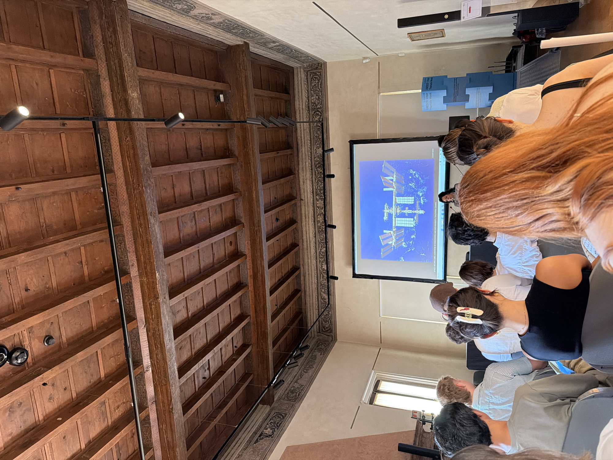 The image shows a room with a high, coffered ceiling, likely in a historical building in Mantua, Italy. The ceiling is made of wood and adorned with intricate carvings, with a track lighting system installed. The walls have decorative borders near the ceiling, showcasing ornate patterns. A large screen projects an image of the International Space Station (ISS). Several people are seated, facing the screen, seemingly attending a lecture or presentation. Many people are visible and appear to be focused on the presentation. Some individuals have their backs to the camera, revealing their hairstyles and attire. The room is well-lit, with additional light coming in from a window. Overall, the scene suggests an educational or informative event taking place in a historically rich architectural space.