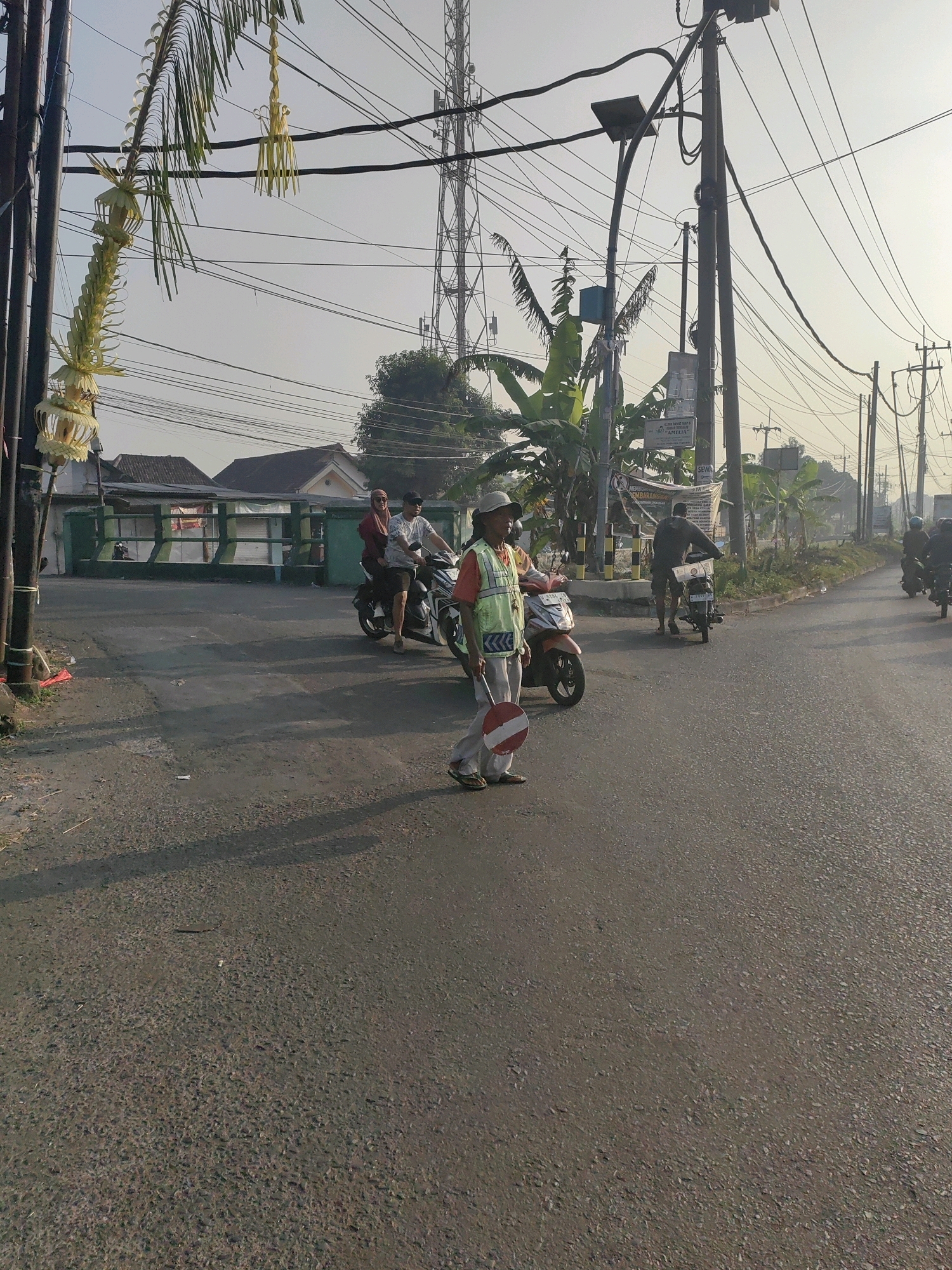 In Keboananom, Indonesia, on a paved road during daylight, a person wearing a reflective vest and hat stands in the foreground holding a red and white stop sign. They are positioned on the right side of the road. Behind them, on the left, a motorcycle with two occupants is stopped. Further down the road, several other motorcycles are visible, some with riders. Utility poles and numerous overhead wires dominate the background, with some greenery and buildings also present. The sky is bright and appears hazy.
