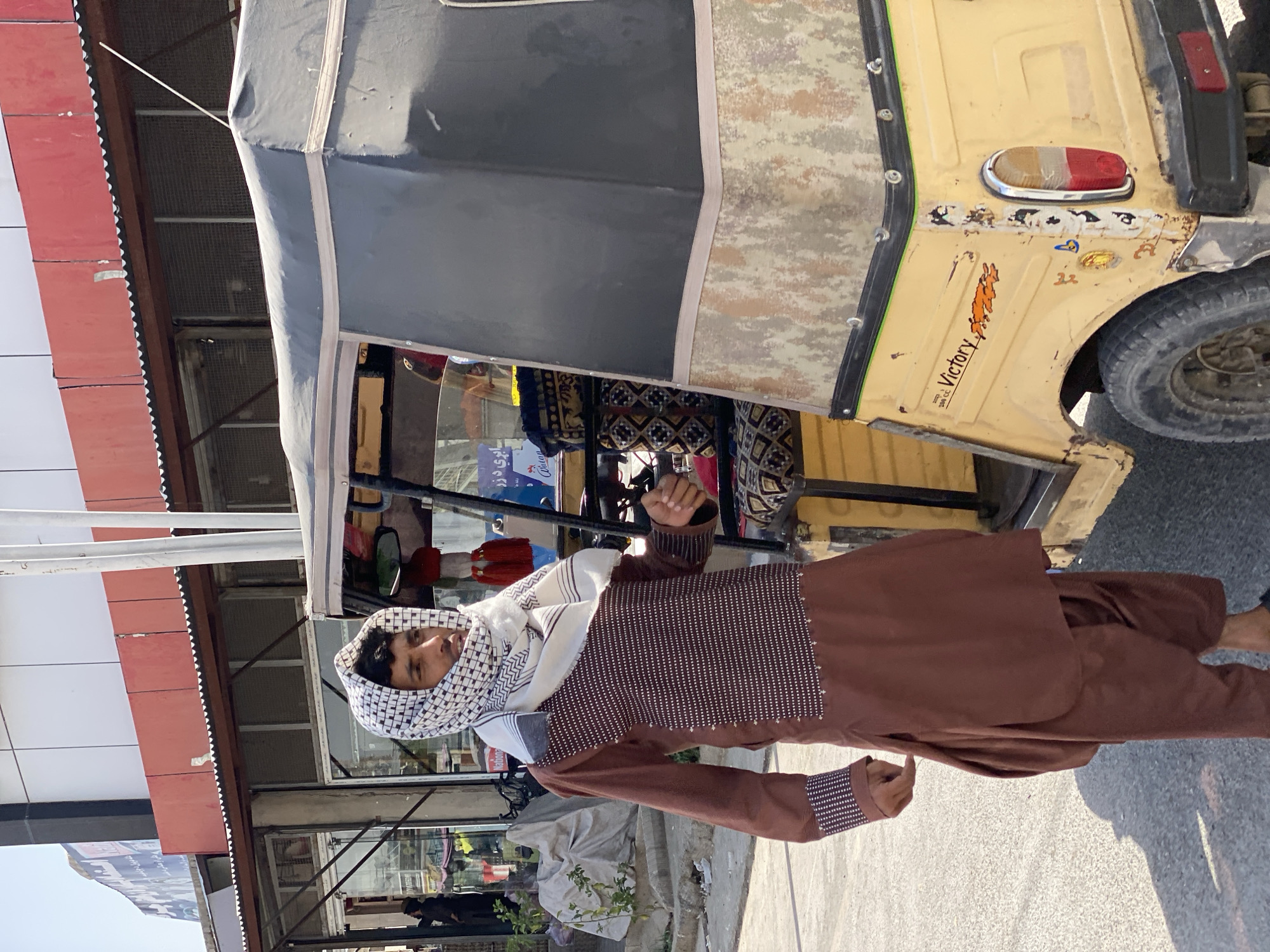 A man wearing a brown shalwar kameez and a checkered keffiyeh is standing next to a yellow auto-rickshaw on a street in Kandahar, Afghanistan. The auto-rickshaw has a dark grey canopy and is decorated with decals, including one that reads "Victory 250 CC". The man is looking to his right, with his right hand slightly extended. Behind him, there are shops with red awnings and white facades. The ground is paved and a shadow of the man is cast on it. The sky is clear and blue.
