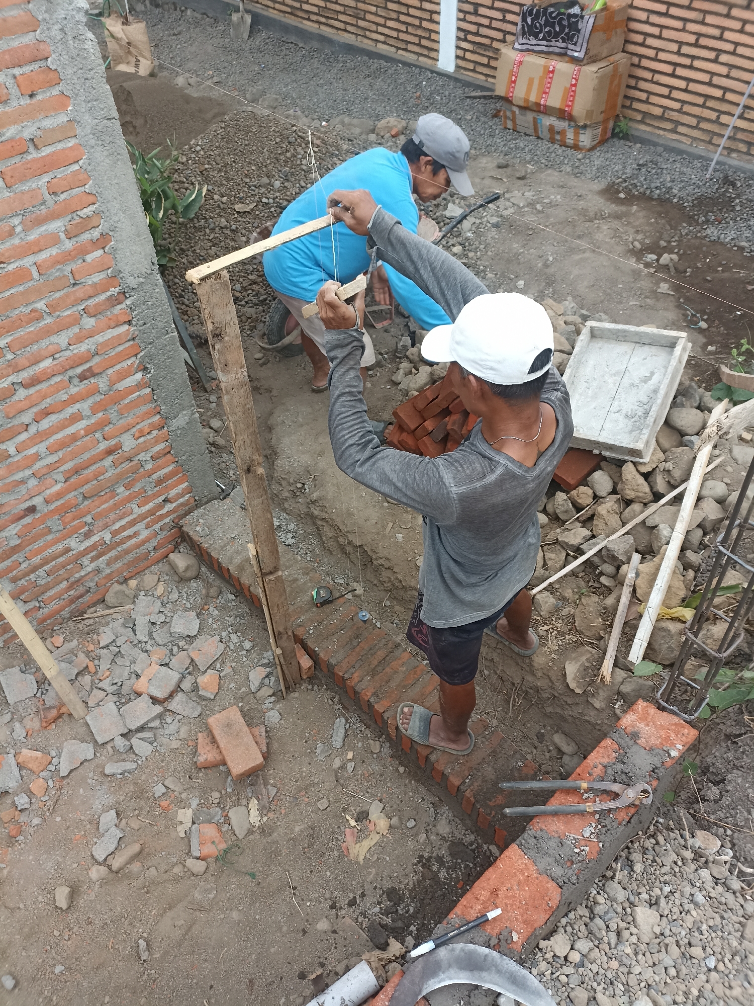 In Mojopanggung, Indonesia, two construction workers are engaged in bricklaying. The scene depicts an outdoor construction site with visible brick walls in various stages of completion. One worker, wearing a blue shirt and a grey cap, is bent over, seemingly assisting with the alignment of bricks. The other worker, in a grey long-sleeved shirt and black shorts, is wearing a white cap and is holding a wooden plank horizontally, with a string attached, likely to ensure precise alignment during the bricklaying process. Numerous red bricks, piles of gravel, and construction debris are scattered around the work area. Tools such as pliers and a tape measure are also visible. The overall environment suggests ongoing building activity in a somewhat rugged outdoor setting.