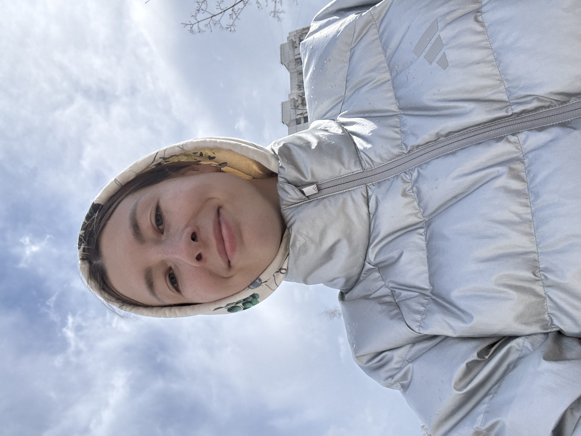 A low-angle, close-up selfie depicts a young woman wearing a silver puffer jacket with the hood up, against a backdrop of a cloudy sky. The hood has a patterned lining. In the upper portion of the frame, bare tree branches are visible, and partially obscured by the hood. In the background there is a partially visible building, which is suggestive of Moscow's urban architectural style. The sky has a mix of blue and white clouds, indicating daylight conditions.