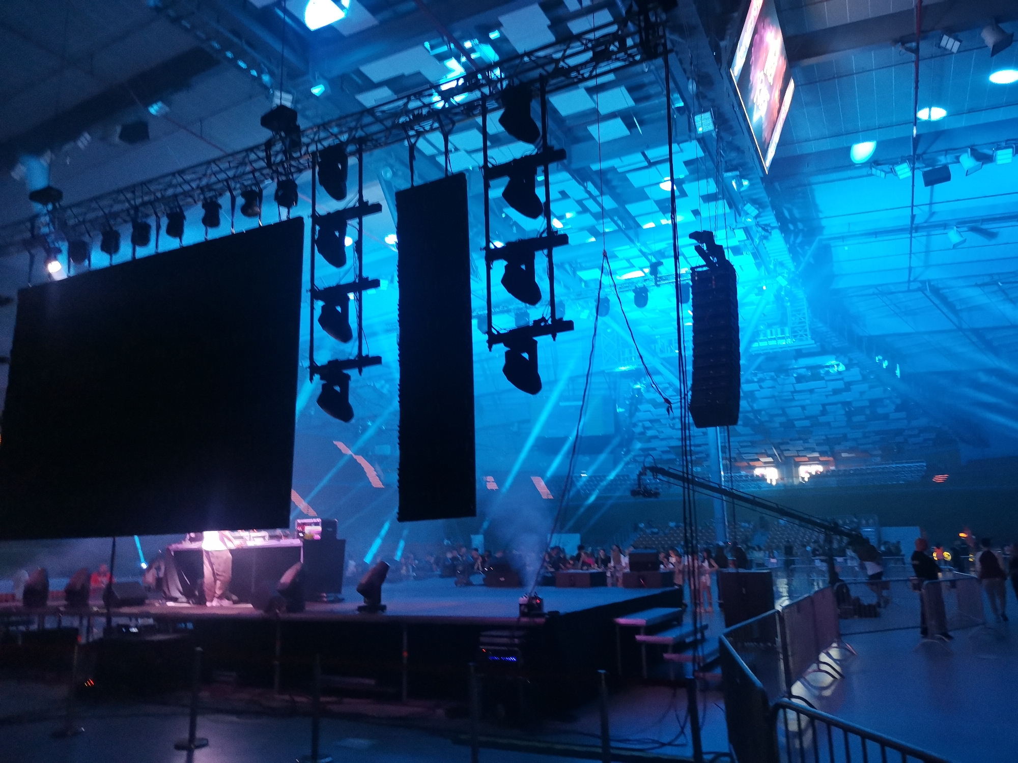 The image captures an indoor event held within a large multi-purpose arena, specifically identified as the Kolodruma (Multipurpose Sports Hall) in Plovdiv, Bulgaria. The scene is illuminated predominantly by a cool blue light originating from numerous stage lights and spotlights, with distinct light beams cutting through a visible atmospheric haze. A central stage is set up, featuring a prominent DJ booth where a person is partially visible behind the equipment. Several stage lights are positioned on the stage floor, and a fog machine emits a plume of smoke. Overhead, multiple metallic trusses suspend large, dark rectangular screens, vertical speaker arrays, and various lighting fixtures. To the left, a wide, dark screen hangs vertically, accompanied by a narrower, equally dark screen to its right. Further right, tall, vertical speaker stacks are suspended. In the upper right background, a large screen displays brightly lit graphics. A camera crane extends from the right side towards the stage area. Metal barriers delineate the space in front of the stage from the audience. Numerous individuals are visible in front of and around the stage, some standing and others seated, observing the event. Several people, likely staff or security personnel, are positioned near the barriers on the right. The arena's high ceiling features a patterned, tiled structure with integrated lighting fixtures, and seating tiers are visible in the distant background.
