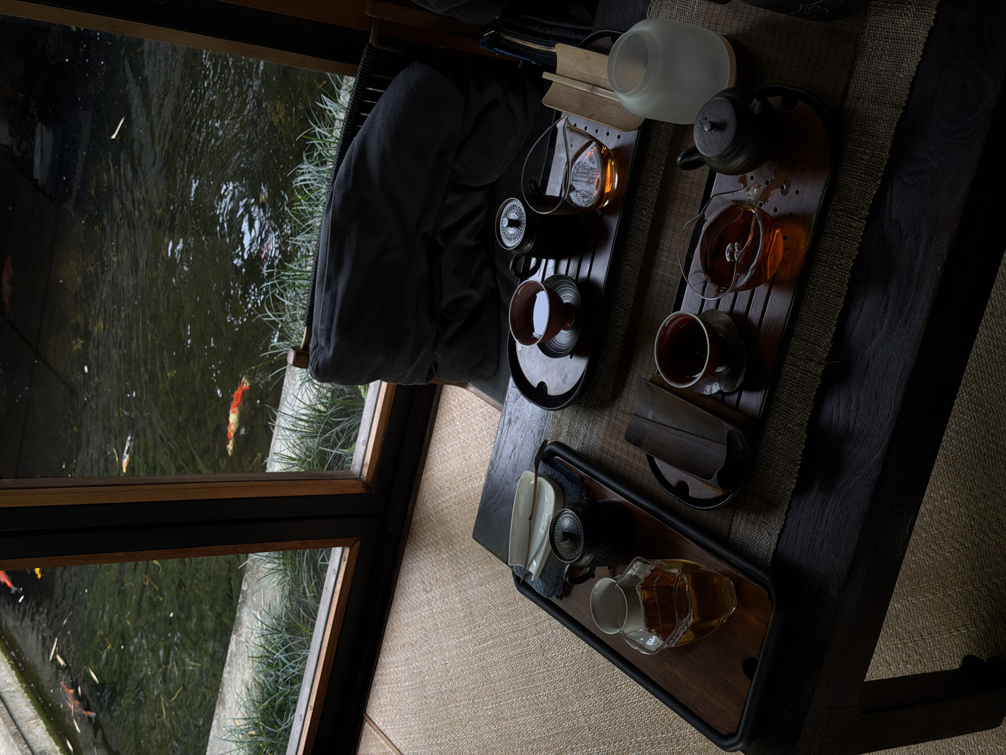 The scene depicts an indoor setting, likely a tea house or residence, in Singakerta, Indonesia, offering a view of an outdoor pond. In the foreground, a dark wooden table is positioned on a woven mat floor. Two separate tea sets are arranged on the table, each on dark wooden trays. The set on the left features a clear polygonal glass pitcher filled with amber liquid, a small black teapot, and two white ceramic shallow dishes with a dark utensil. The set on the right includes a dark red ceramic teacup on a saucer containing dark amber liquid, a small black teapot, a clear glass pitcher with amber liquid, and a light-colored frosted bottle or diffuser. A dark wicker chair with a substantial dark grey cushion is partially visible behind the table on the right. Through a dark wooden-framed window occupying the upper half of the image, a tranquil pond is visible. The pond water is dark green, and several koi fish, identifiable by their orange, white, black, and red/white patterns, swim in it. Green aquatic vegetation or grass lines the concrete edge of the pond. No individuals are present in the image.