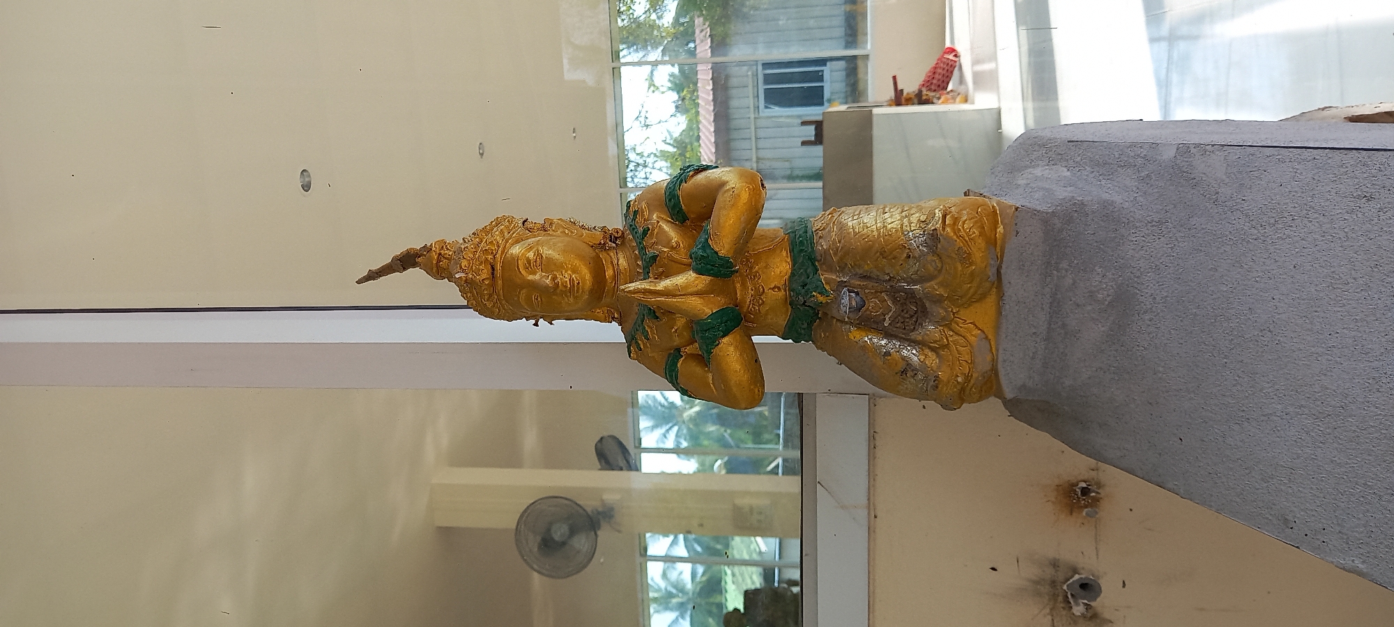 The image shows a golden Buddha statue with green accents. The statue appears aged and is mounted on a grey, concrete base. The setting appears to be indoors, possibly a temple or religious building, as evidenced by the reflection in the glass behind the statue, which shows a window and other structures. The room's walls are off-white. There are visible imperfections and repairs on the wall behind the statue. In the reflection, a fan can be seen mounted on the wall. The location of the image is Moo 4, Thailand, based on the geographic coordinates provided.