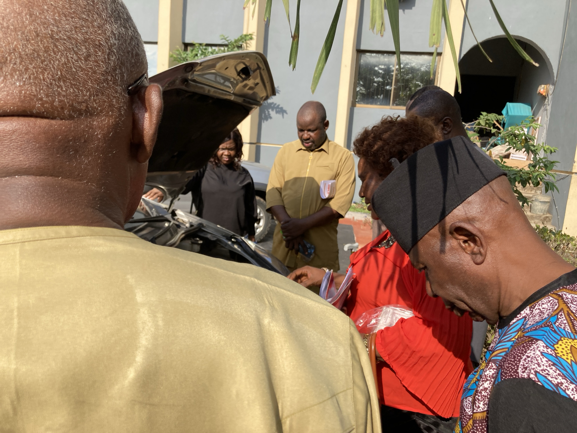 The image depicts a group of several people gathered outdoors around the open hood of a car. In the immediate foreground, an older man with short, graying hair, wearing a light-brown/khaki shirt, is seen from behind. To his right, a man in profile wears a black cap and a colorful patterned shirt with blue, brown, and yellow motifs, looking downwards.
In the midground, a man in a mustard yellow collared shirt with a receding hairline and beard looks down towards the car's engine. Next to him, a woman with dark, curly hair, dressed in a red pleated top, also looks down and is holding items in clear plastic bags. Another woman in a black top is partially visible under the raised car hood on the left, also looking into the engine bay. A fifth person, a man, is partially seen on the right, also focused on the car.
The activity suggests a car inspection, repair, or discussion about a car issue. The scene is brightly lit by sunlight, indicating daytime, with clear skies and visible shadows. In the background, a light-colored building with windows and an arched entryway is visible, along with green palm fronds and other plants. No discernible text is visible. The location is Umuahia, Nigeria.