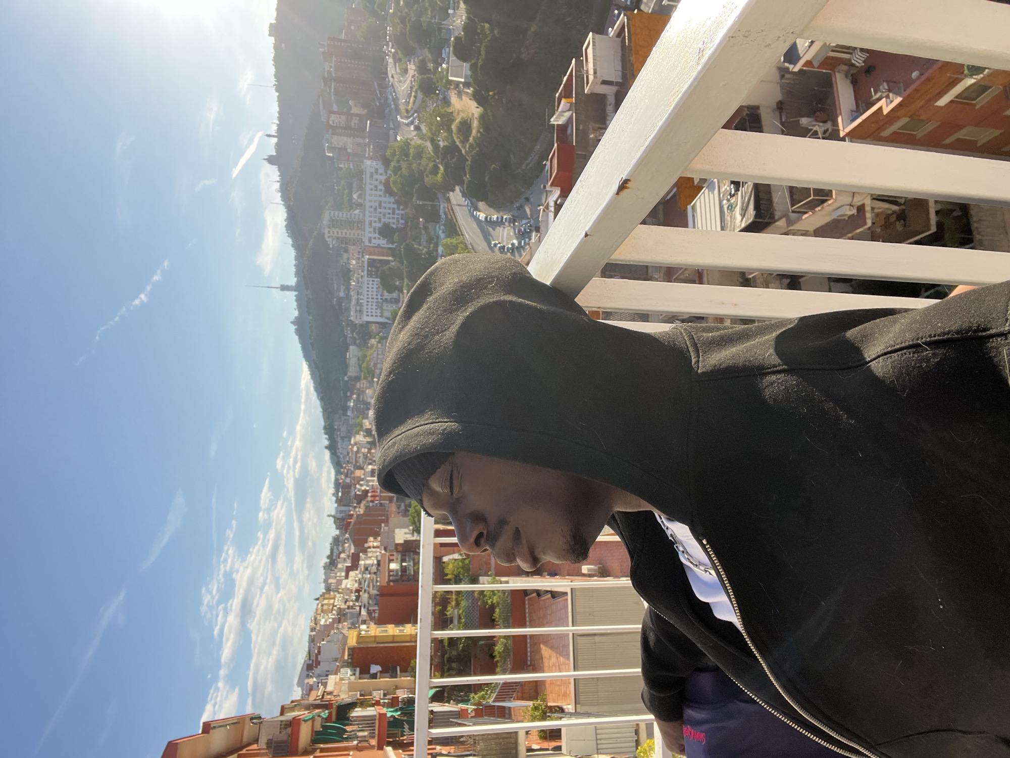 A medium shot shows a person with dark skin and a black hooded sweatshirt, standing on a white railing overlooking the city of Barcelona, Spain. The person is wearing a white shirt underneath the sweatshirt and is looking down. The city of Barcelona is visible in the background, with buildings and streets stretching out to the horizon. The sky is blue with some clouds. The photo is rotated clockwise. The location seems to be taken from an elevated viewpoint, likely a balcony or rooftop.